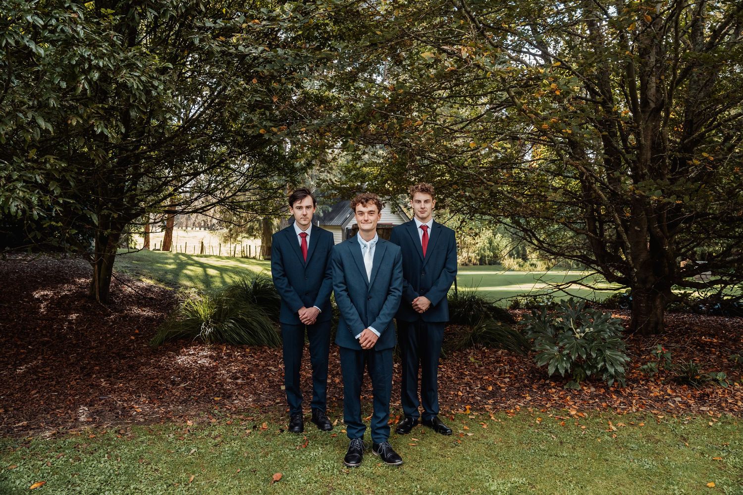 Three young men in navy suits and red ties posing outdoors under lush green trees on a lawn.