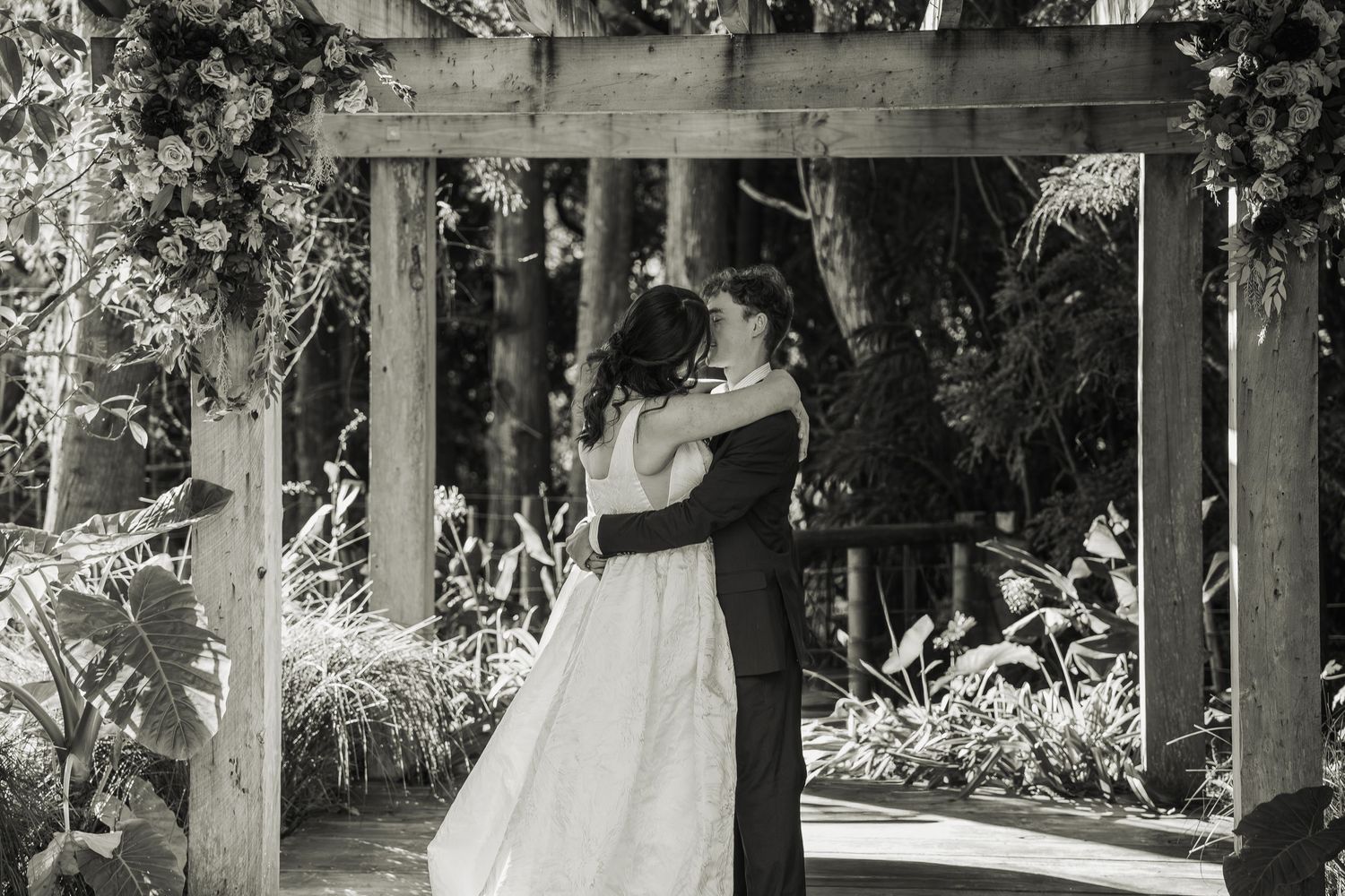 Black and white photo of bride and groom sharing their first kiss under a wooden pergola with floral decorations.