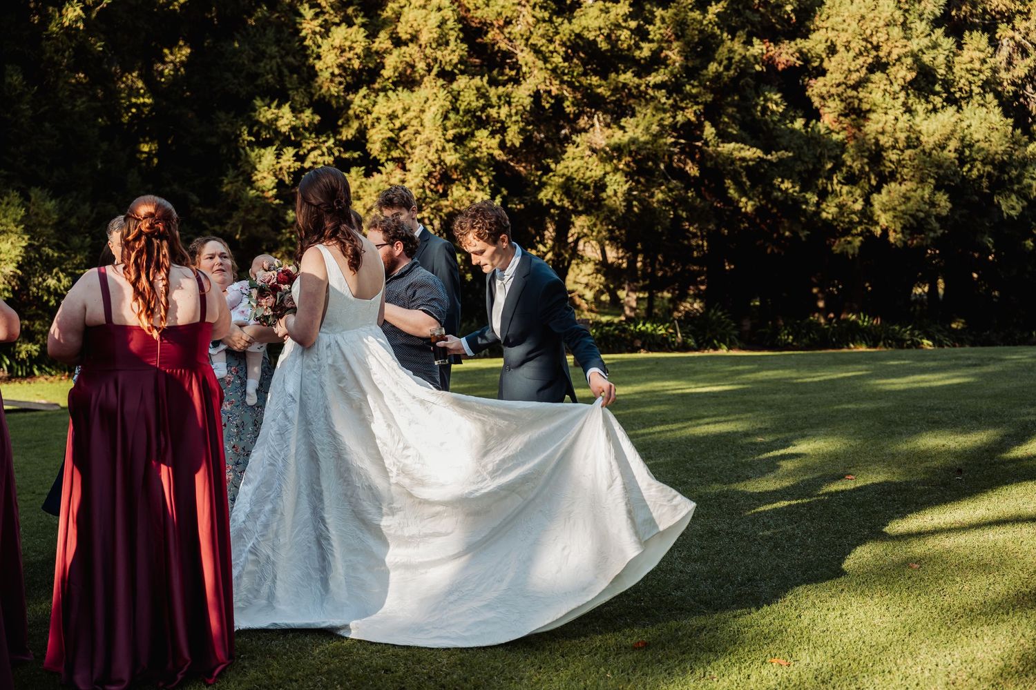 Bridesmaids in burgundy and groom fluffing bride's white lace gown on green lawn.