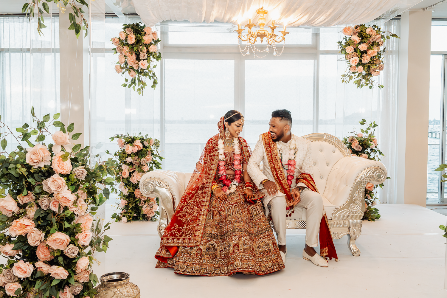 Wedding venue decorated with peach floral arrangements and couple in traditional Indian attire on white sofa.