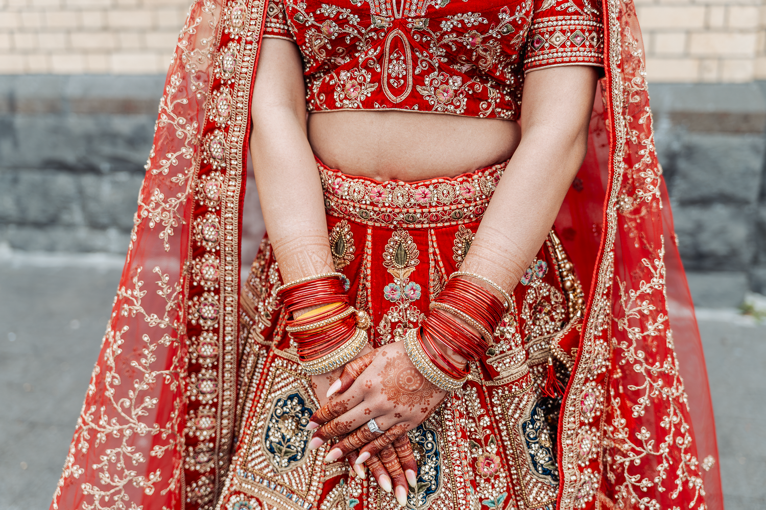Detail shot of henna-adorned hands and red wedding bangles with embroidered lehenga.