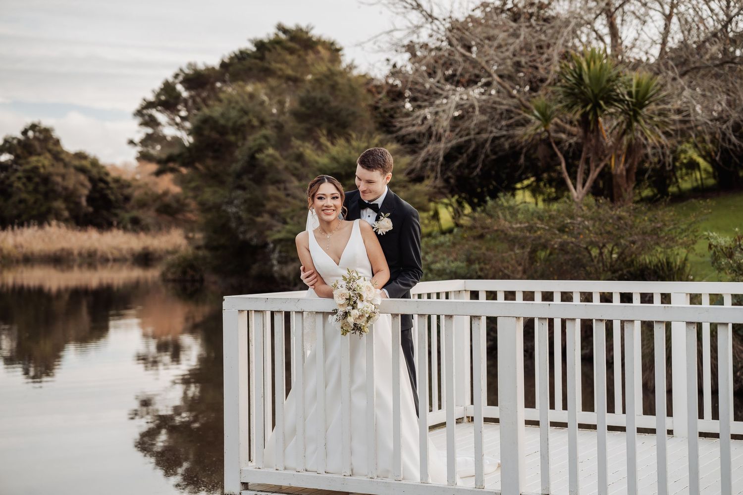A couple stands together on a white wooden deck overlooking a serene pond during sunset.