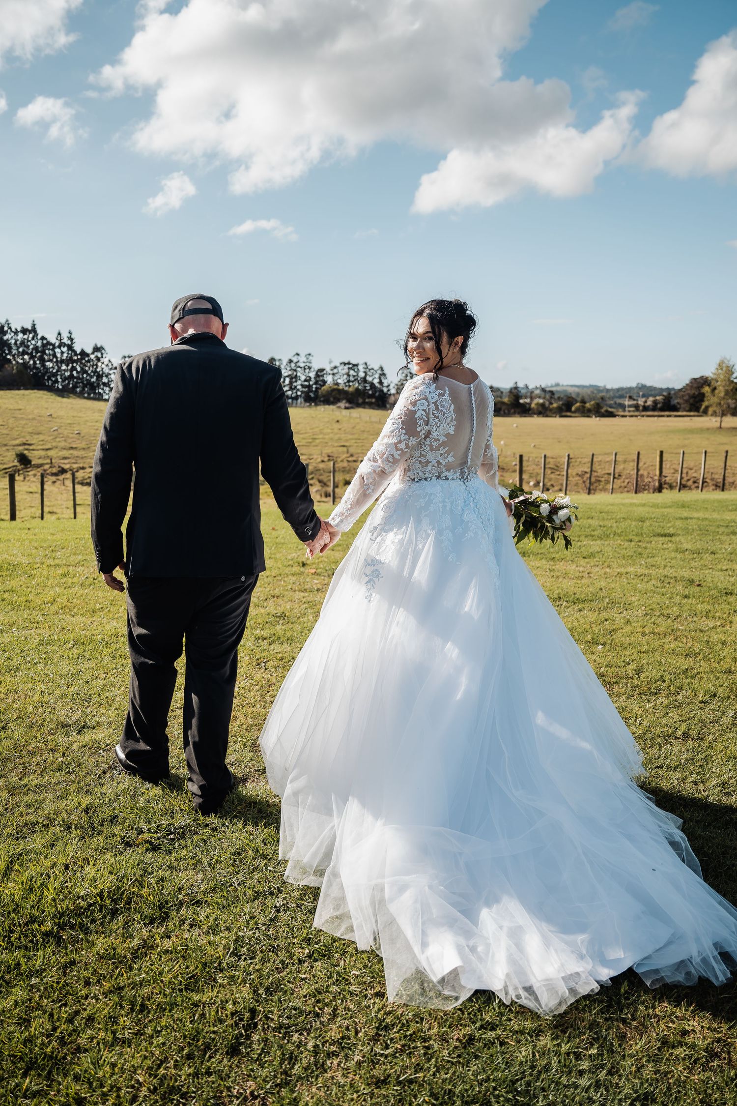 Newlyweds walk hand in hand across green field with bride's lace gown trailing behind.
