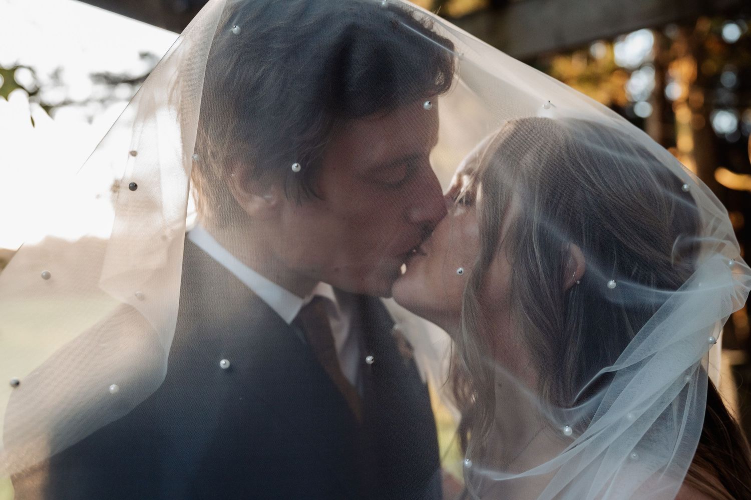 Romantic wedding couple sharing intimate kiss under flowing bridal veil with twinkling lights in background.