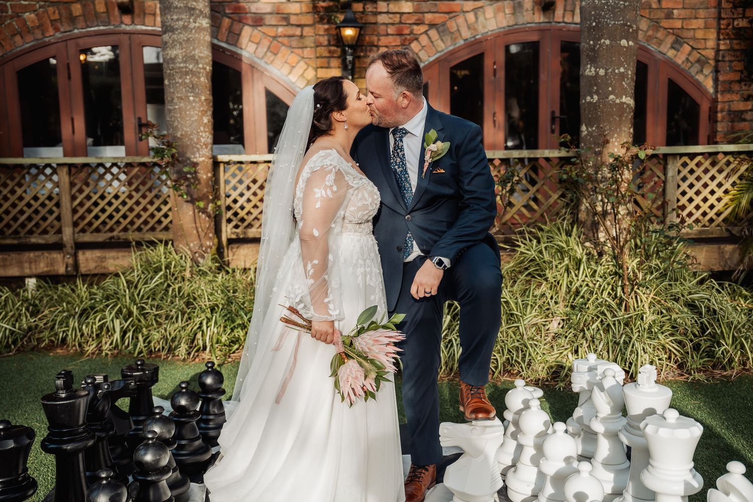 Newlyweds share a romantic moment next to a giant chess set in a garden setting.