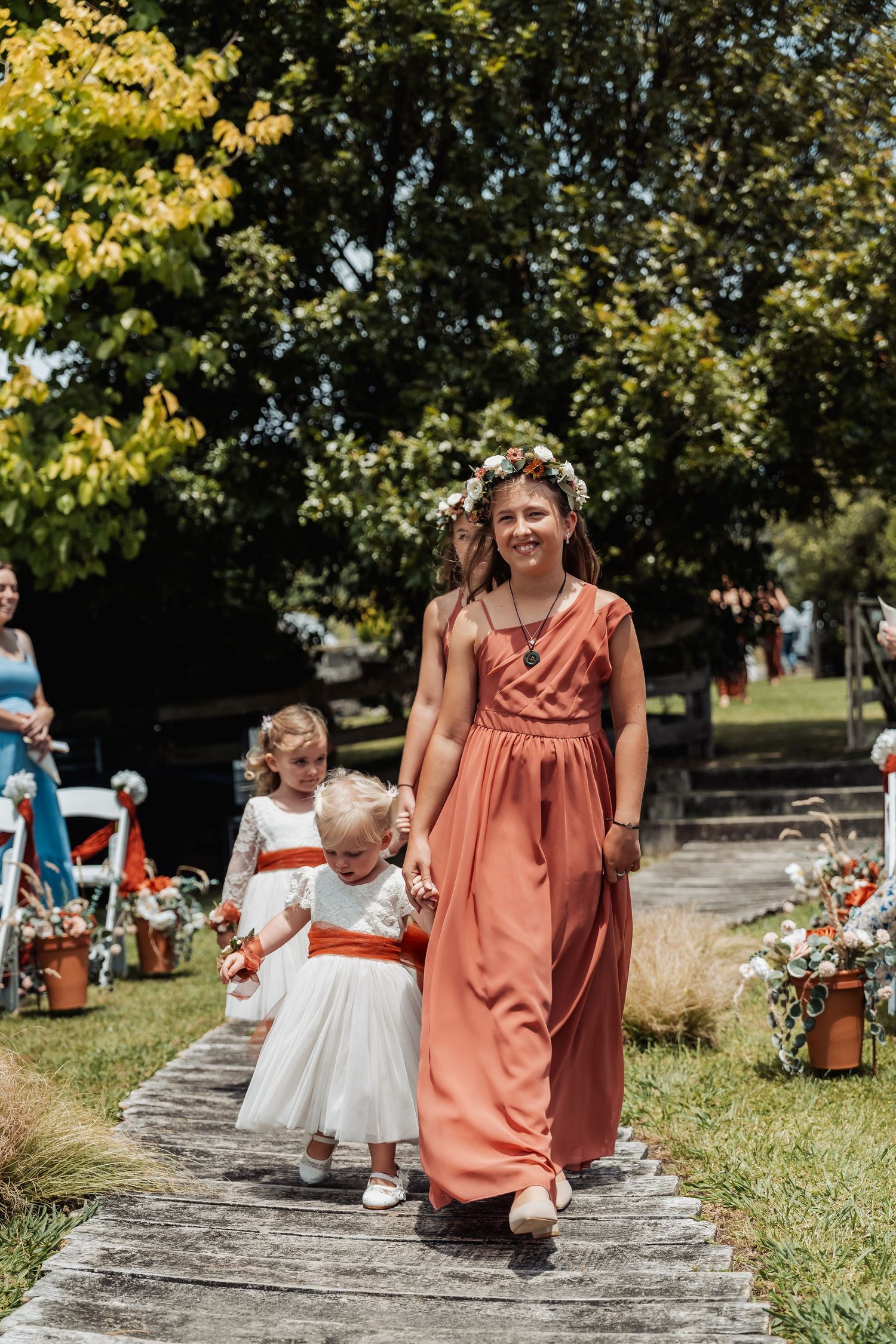 Bridesmaid in coral dress with floral crown leads flower girls down wooden aisle at outdoor wedding.
