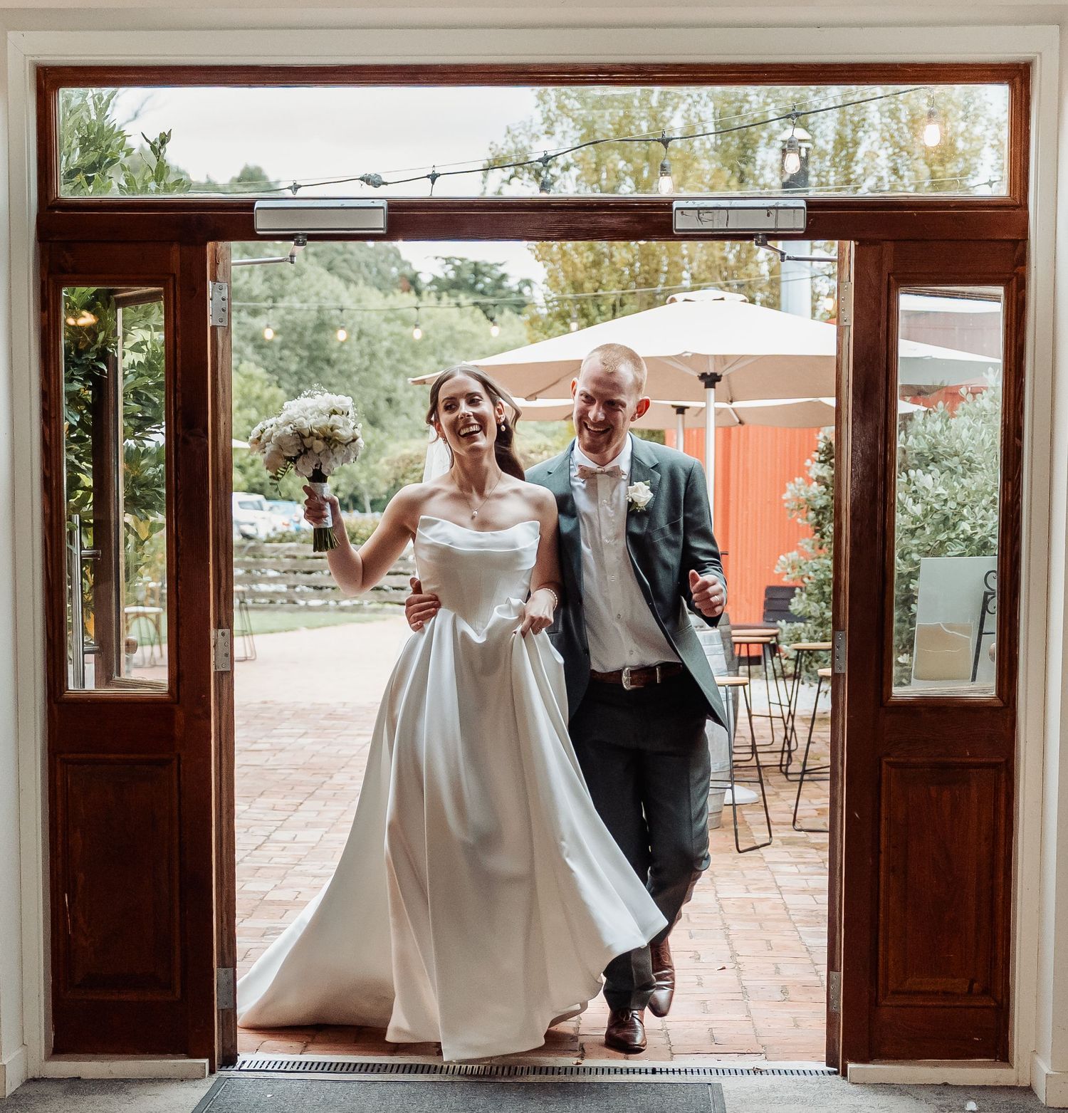 Laughing bride and groom make grand entrance through wooden doors holding bouquet at their wedding reception.
