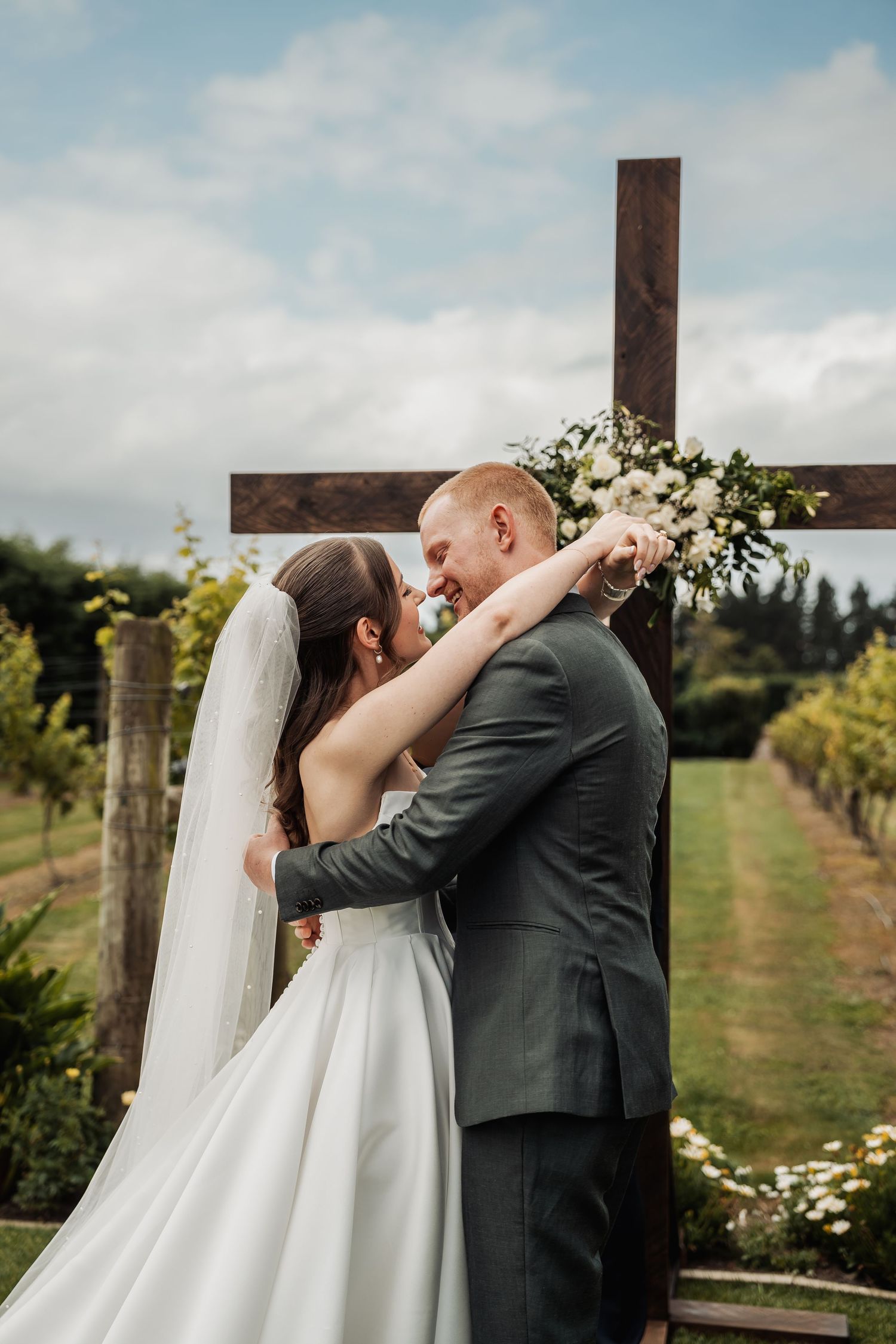 Bride and groom embracing at outdoor wedding ceremony with wooden cross.