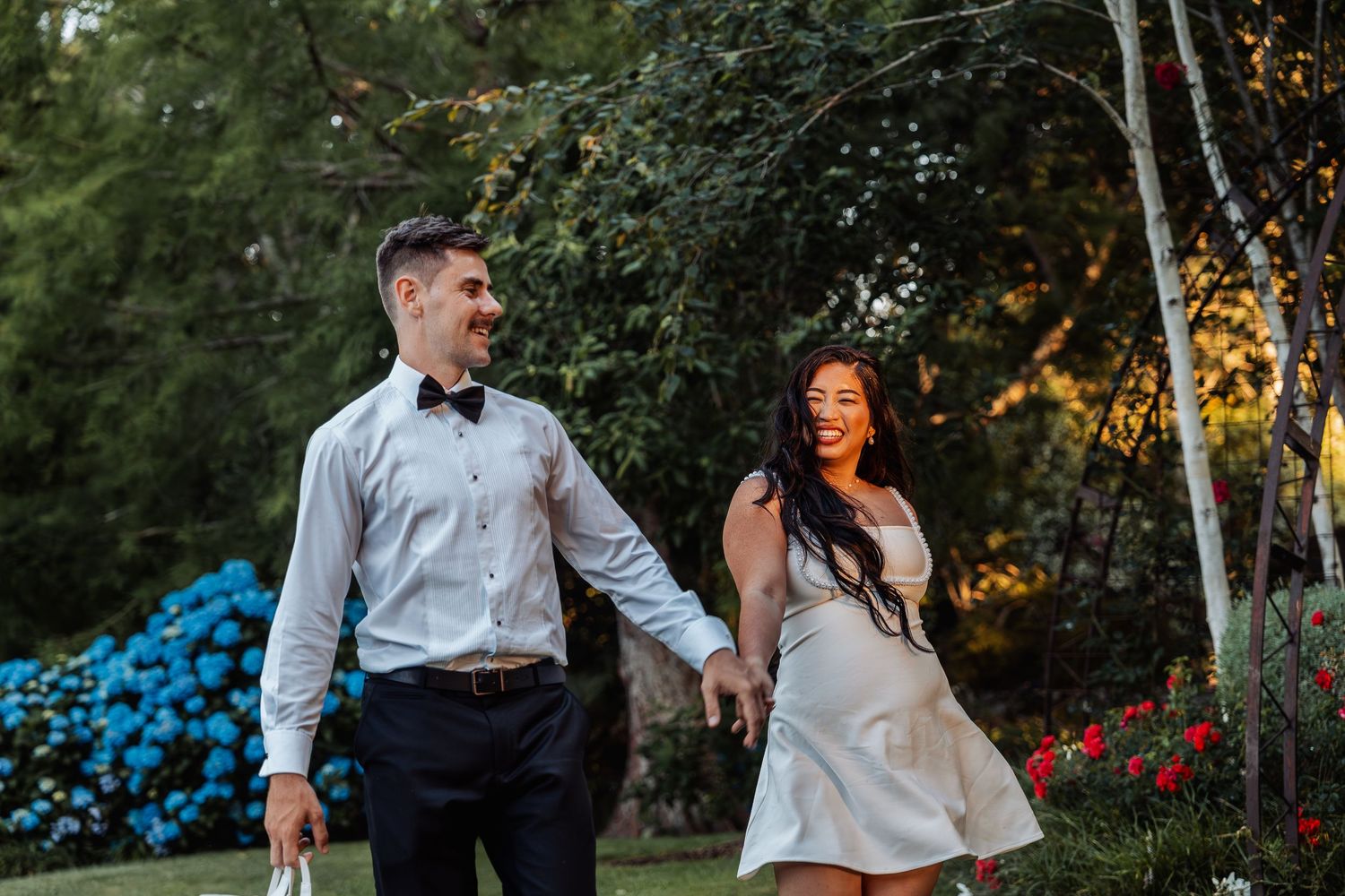 Couple holds hands and smiles while strolling through a colorful garden at sunset.