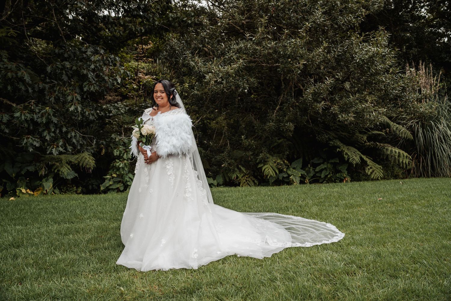 Bride in elegant white off-shoulder gown with fur wrap and long train poses on green lawn.