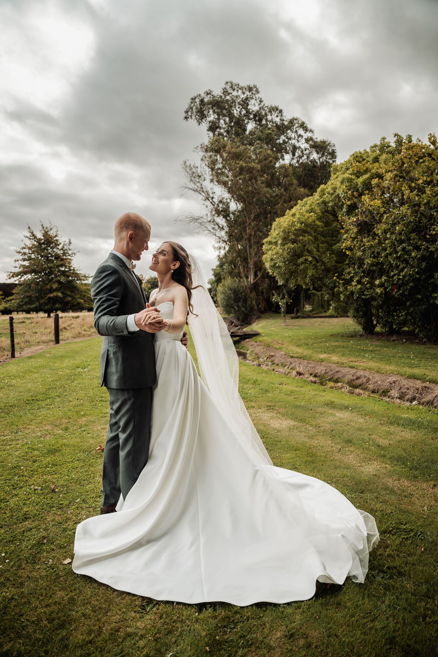 Bride and groom share a romantic moment on lush green lawn, her elegant white gown with long train flowing behind her.