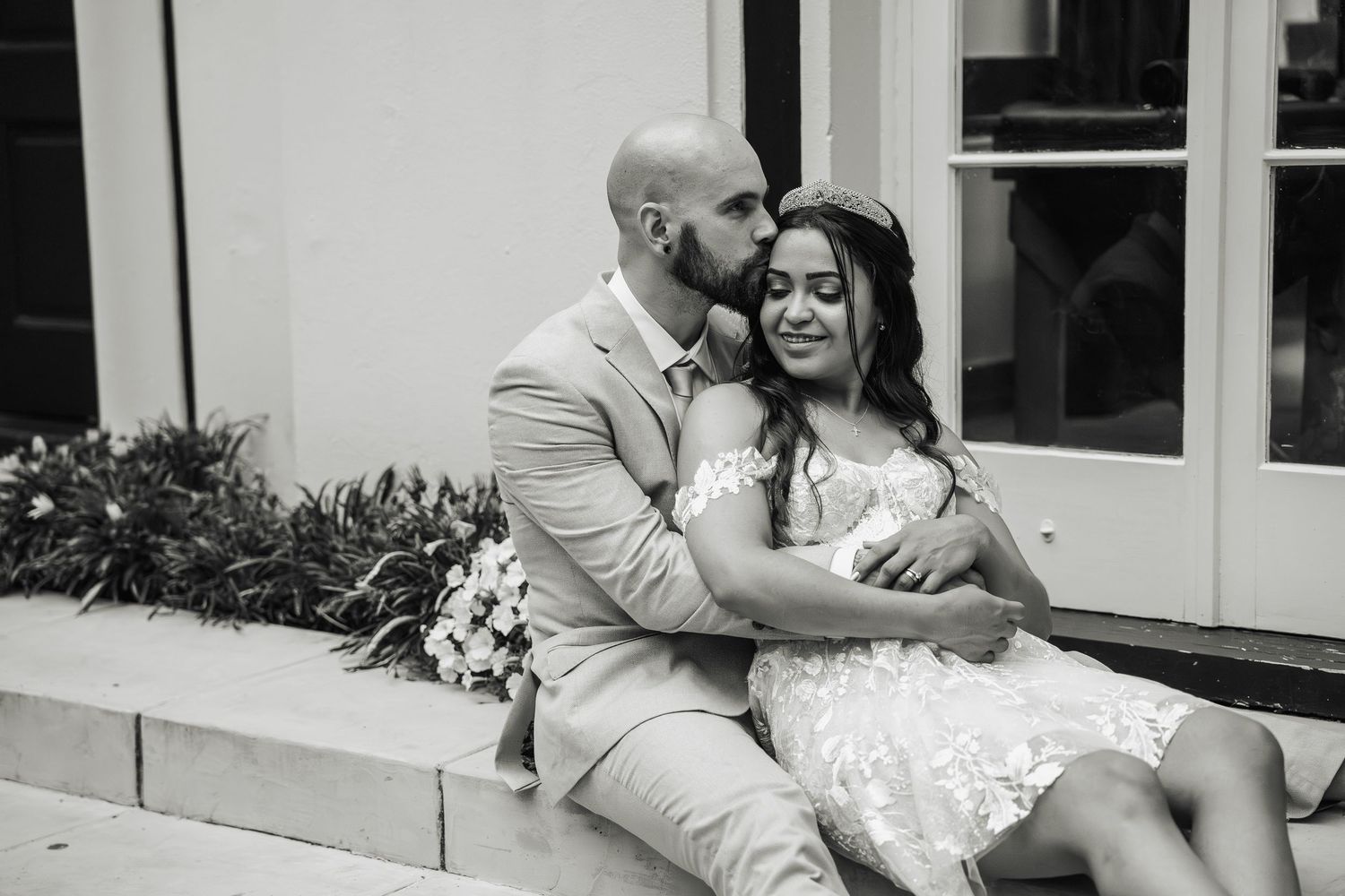 Black and white photo of groom kissing brides cheek as they sit together on outdoor steps with bouquet.