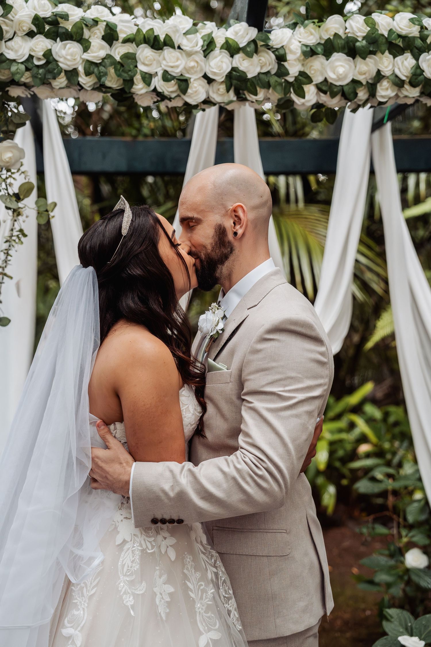 Bride and groom share first kiss under floral arch decorated with white roses and draping.