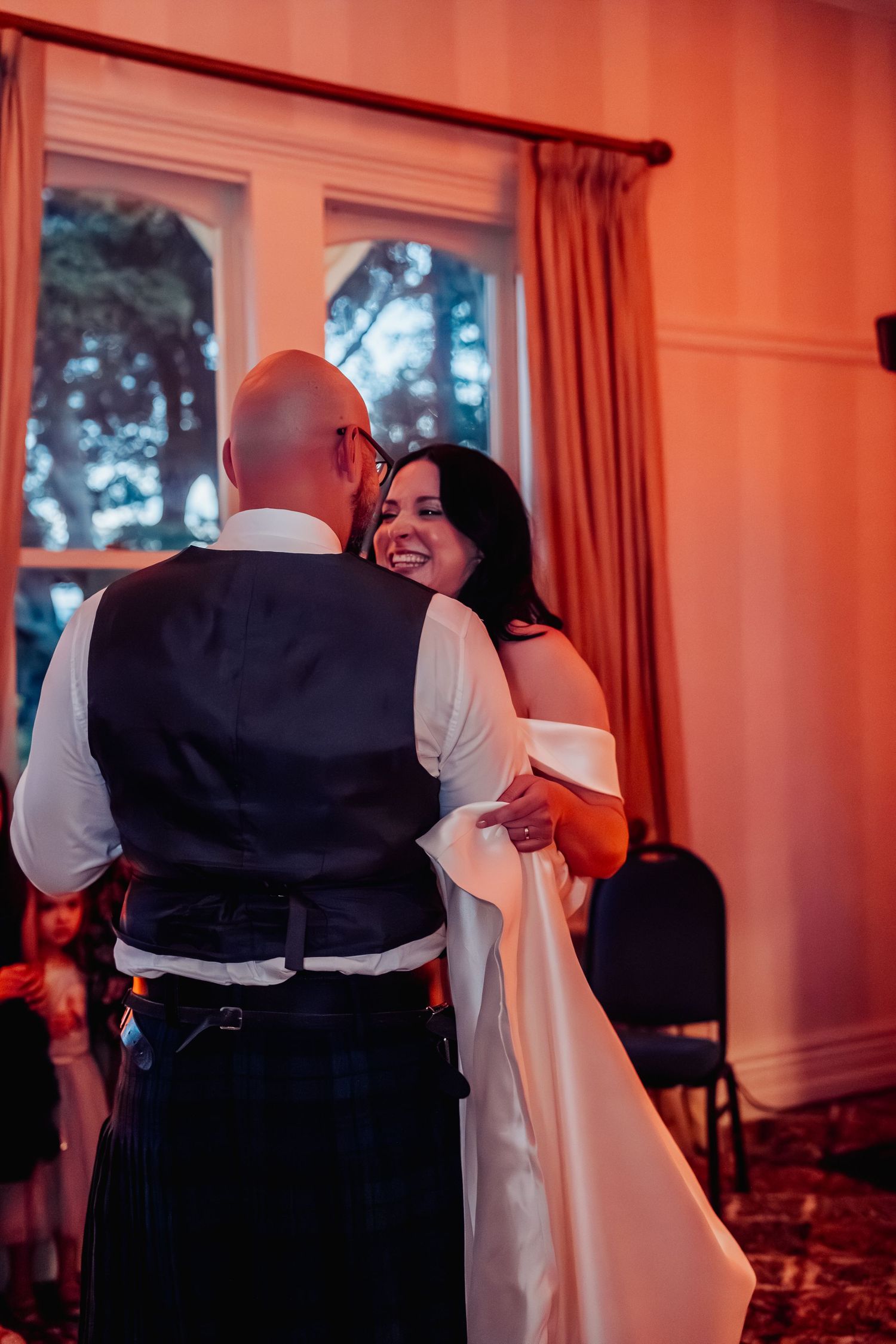 Bride in white gown smiles joyfully while sharing first dance with groom in kilt under warm amber lighting.