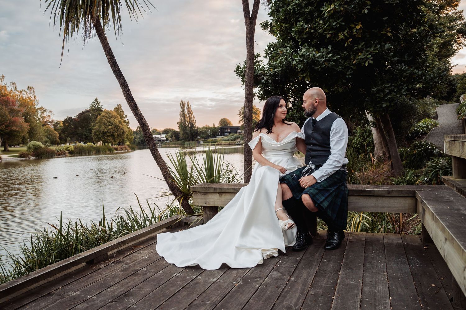 Bride and groom in tartan kilt share a romantic moment seated on a bench by a scenic lakeside at dusk.