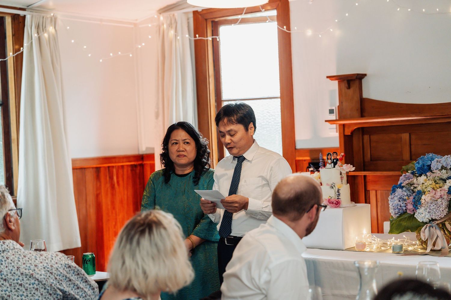 Couple delivers heartfelt speech to wedding guests inside rustic timber reception venue with fairy lights.
