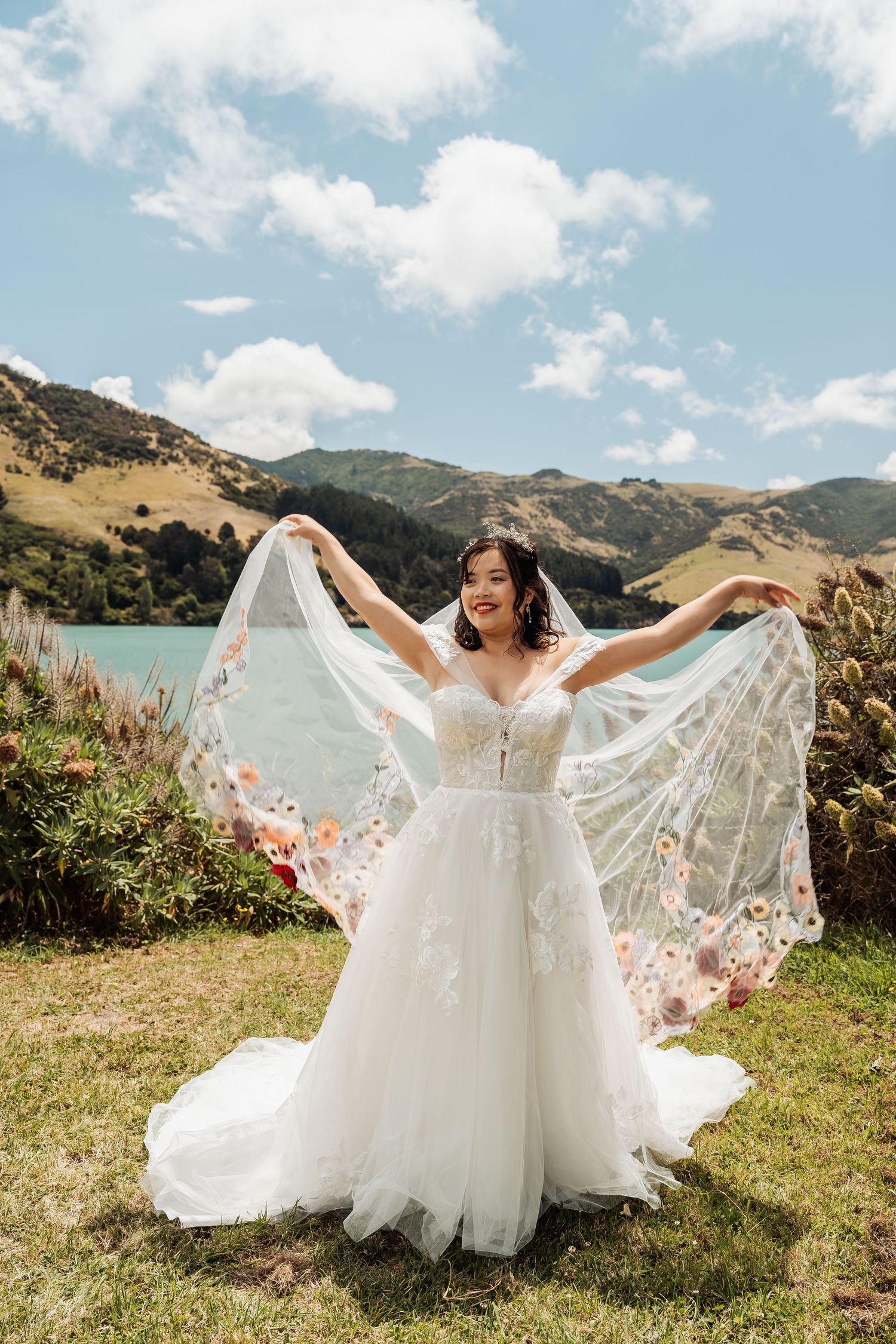 Bride in floral veil and lace gown spreading her veil wide with mountains and lake backdrop on sunny day.
