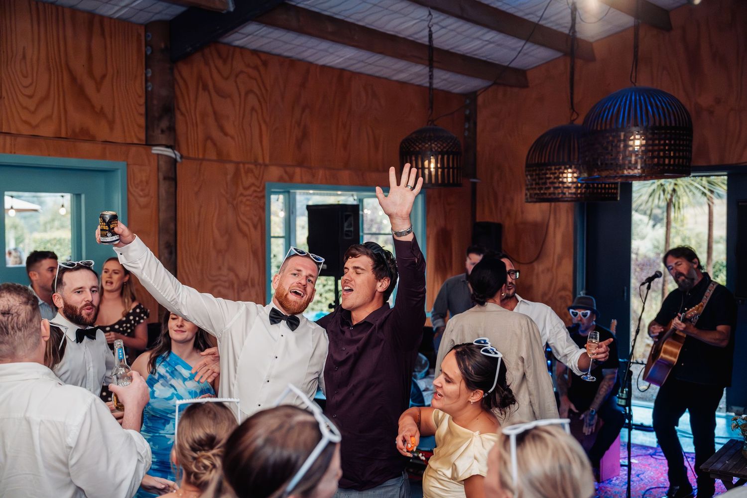Groom and guests celebrating with raised hands on the dance floor at a lively wedding reception.