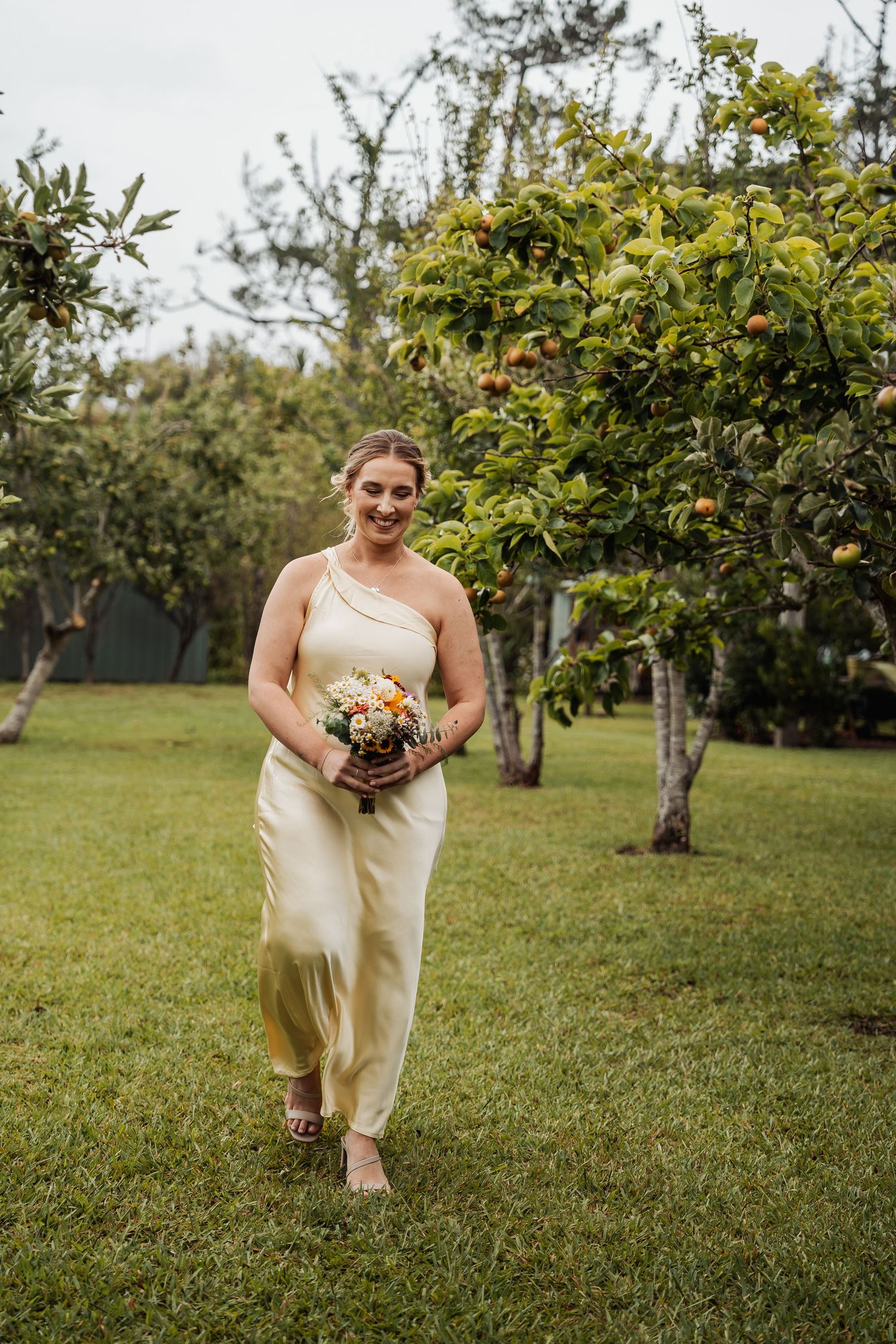 Bridesmaid in yellow one-shoulder jumpsuit walking through garden orchard holding bouquet.