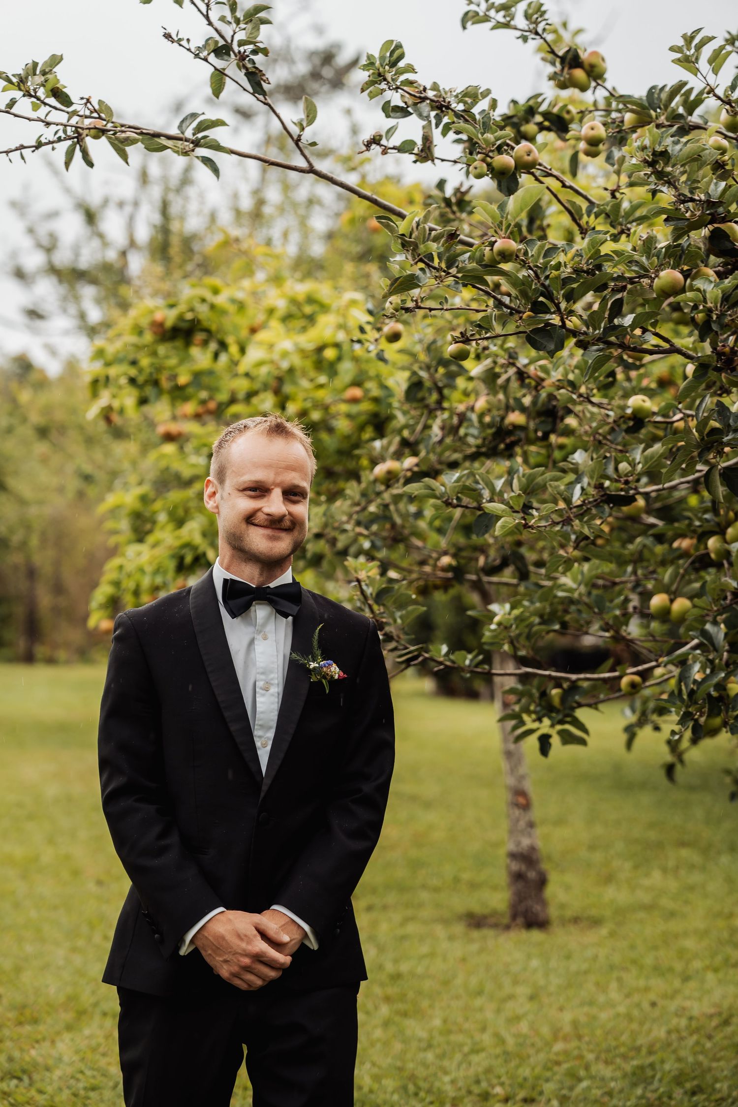Dapper groom in a black tuxedo with bow tie smiling beside a fruit tree outdoors.
