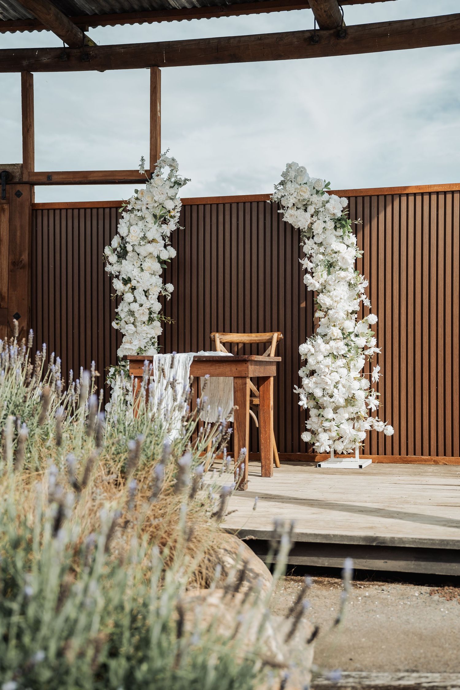 Elegant white floral arch with roses framing a wooden table and chairs on a rustic outdoor wedding ceremony deck.