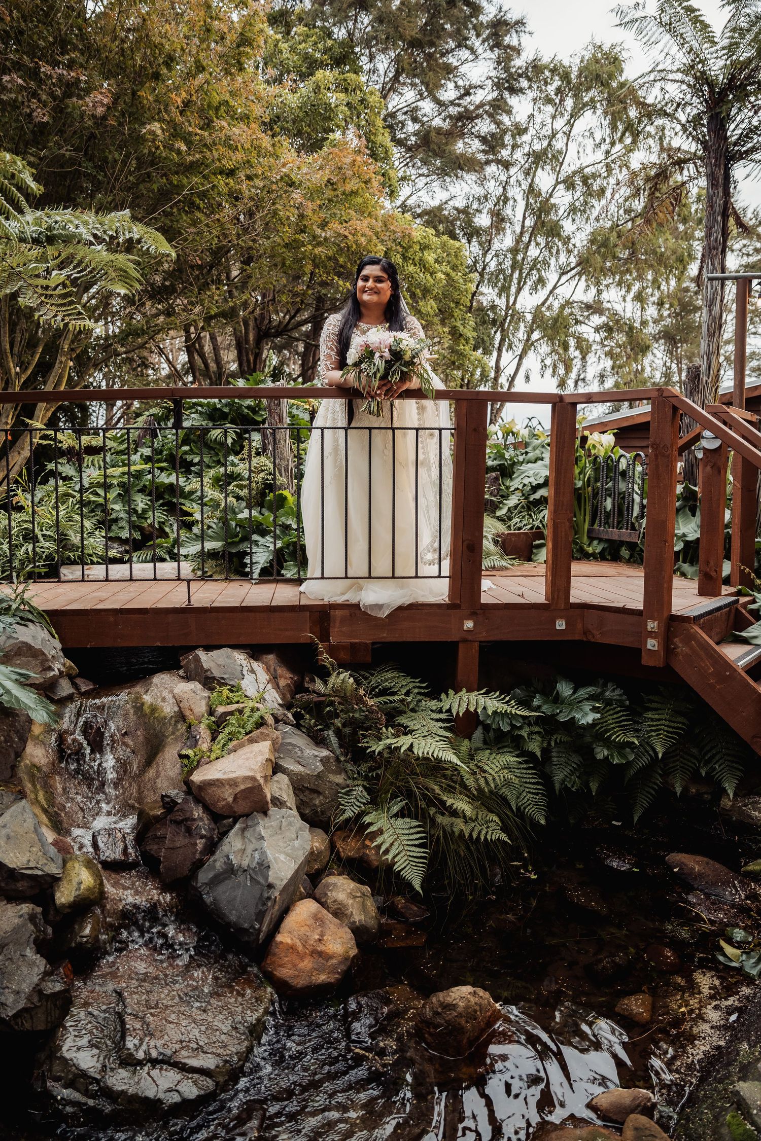 Bride in white gown holding bouquet stands on wooden bridge over garden stream.