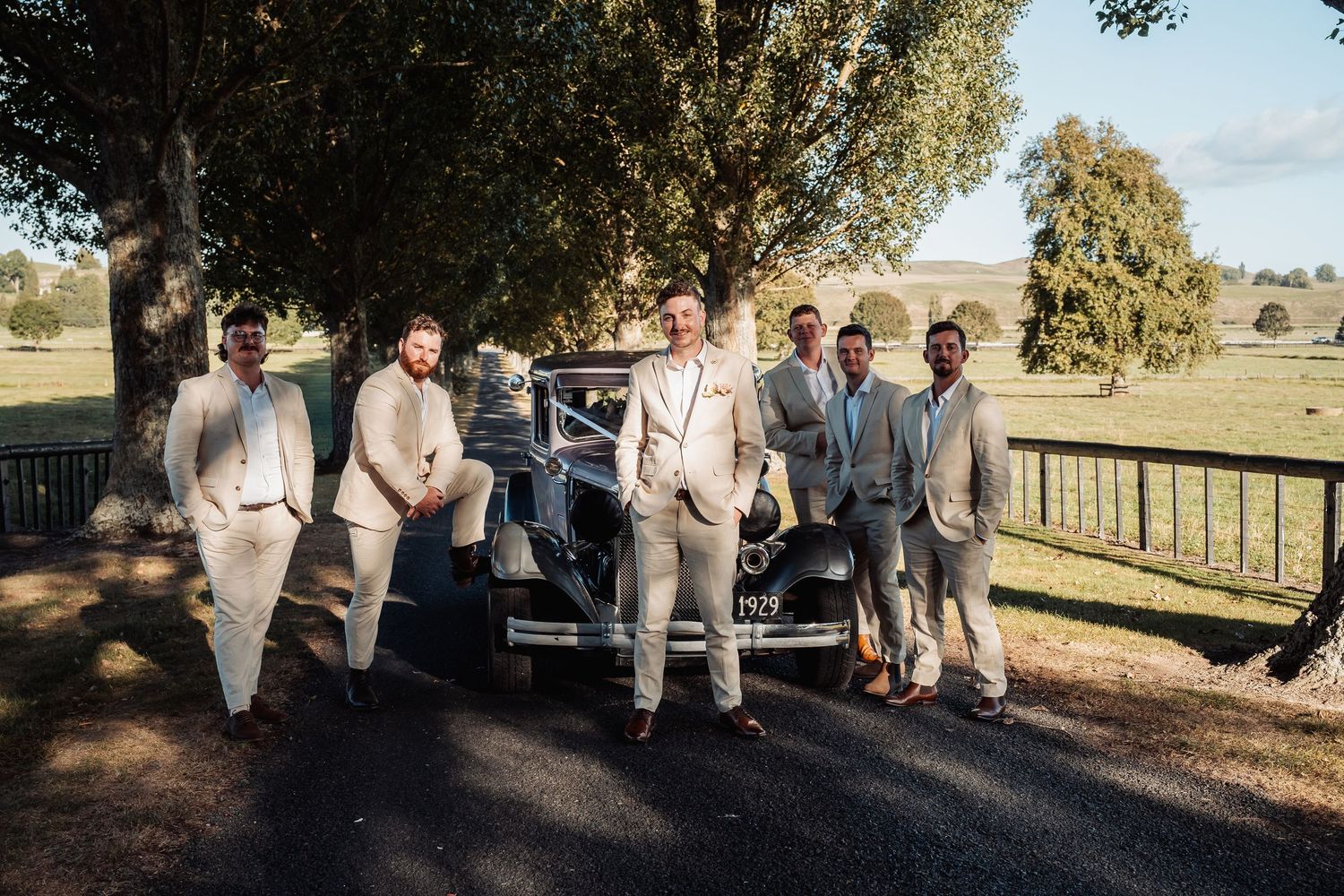Groom and groomsmen in beige suits posing with a vintage car on a sunny day.