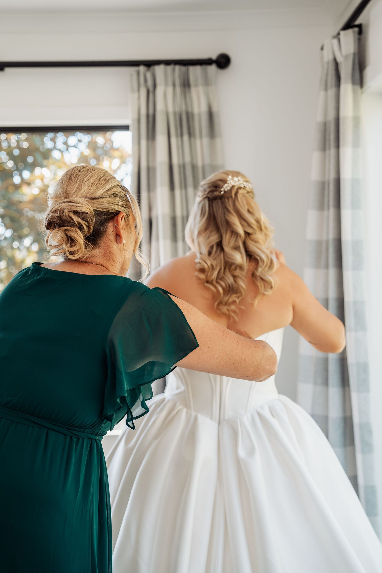 Bridesmaid in green dress helping bride with her white wedding gown near a window indoors.