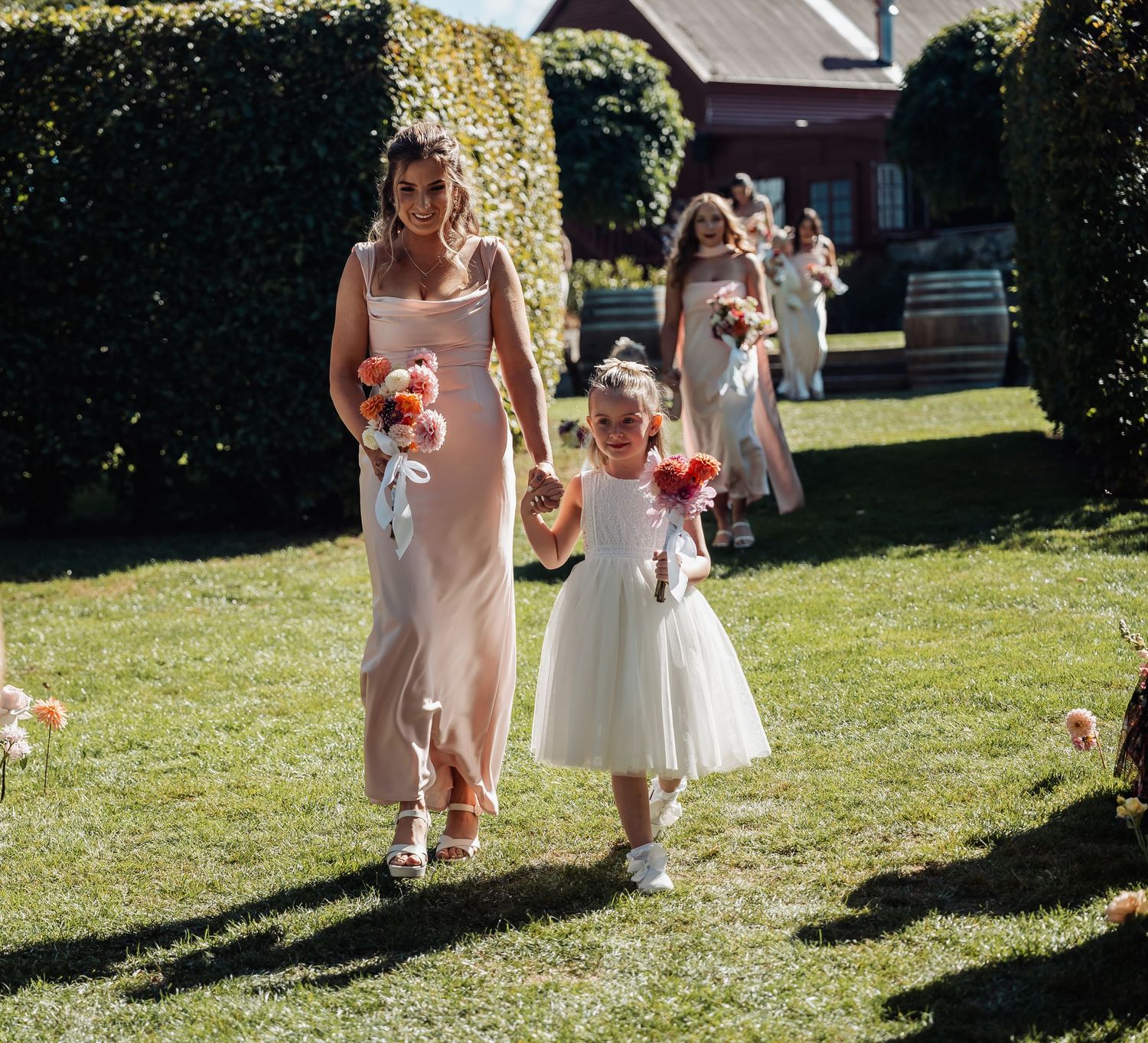 Bridesmaid in blush gown walks flower girl in white dress down outdoor wedding aisle holding coral bouquets.