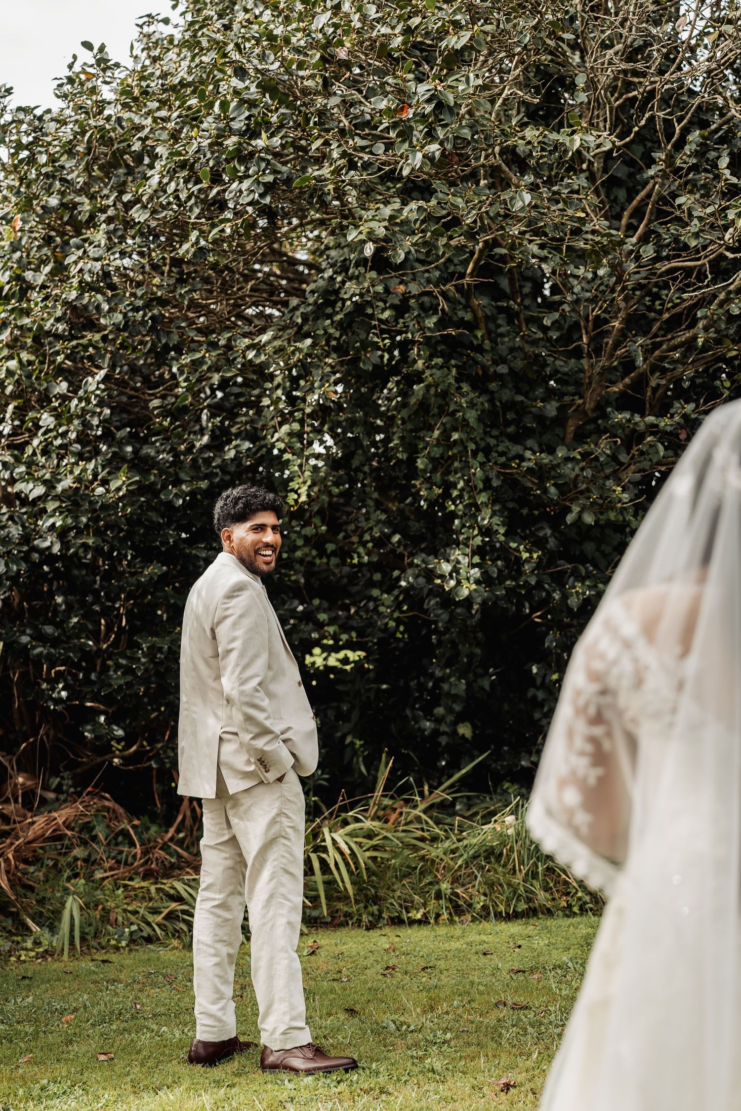 Groom in beige suit smiling at bride during outdoor first look wedding moment with lush green trees.