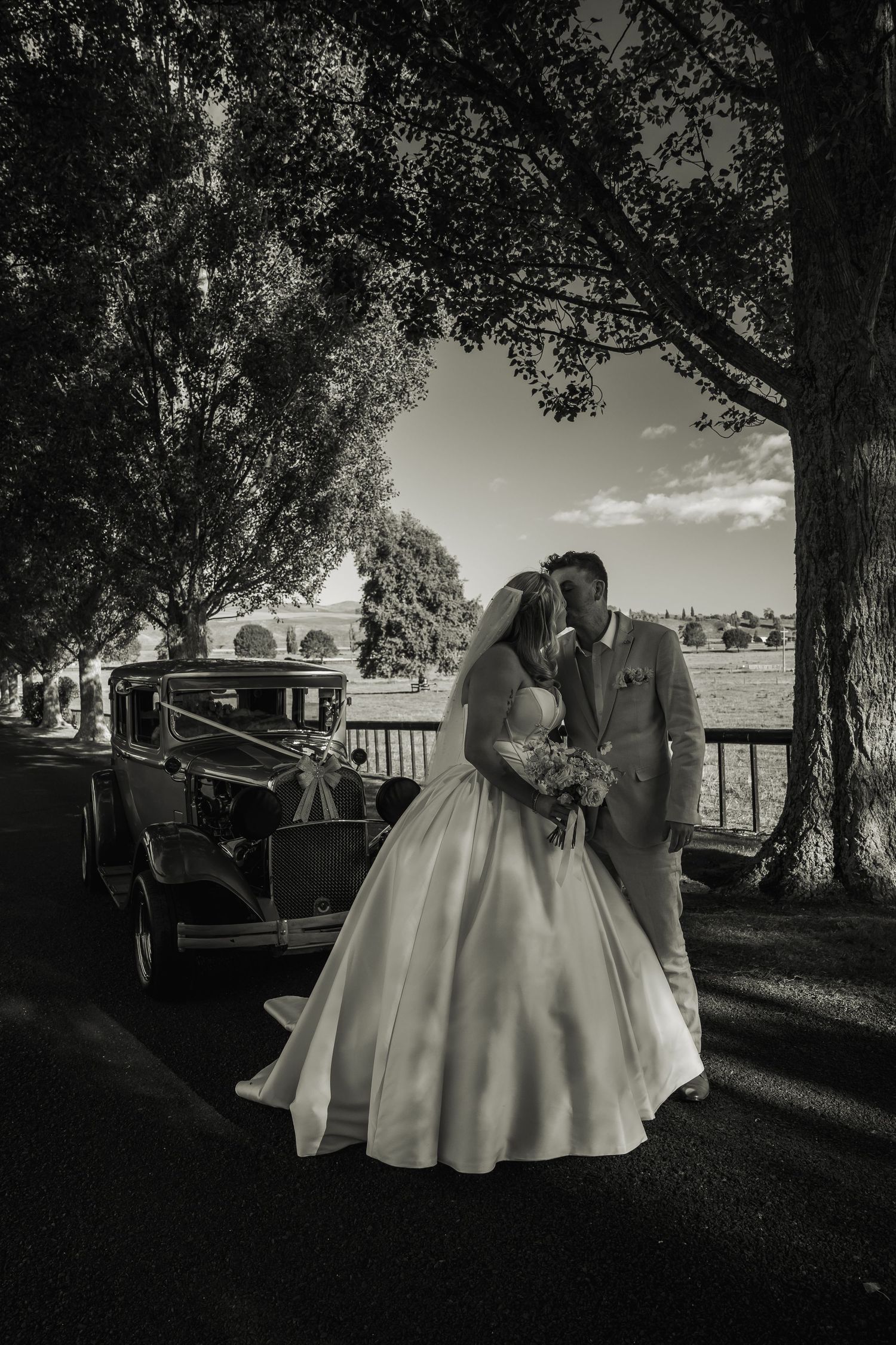 Black and white photo of bride and groom kissing beside vintage car under tree-lined driveway.