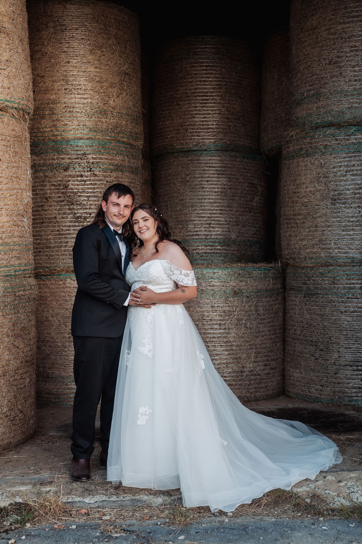 Bride and groom embracing in front of large rustic hay bales in romantic farm wedding portrait.