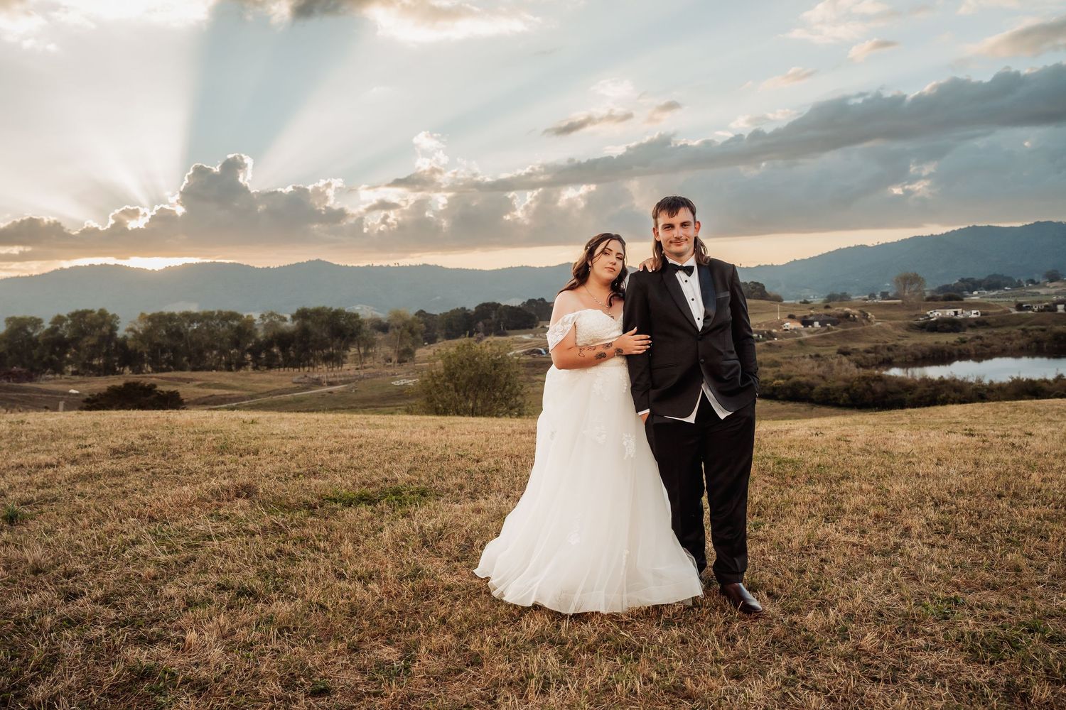 Bride and groom pose together in a field at sunset with scenic mountain and lake views behind them.