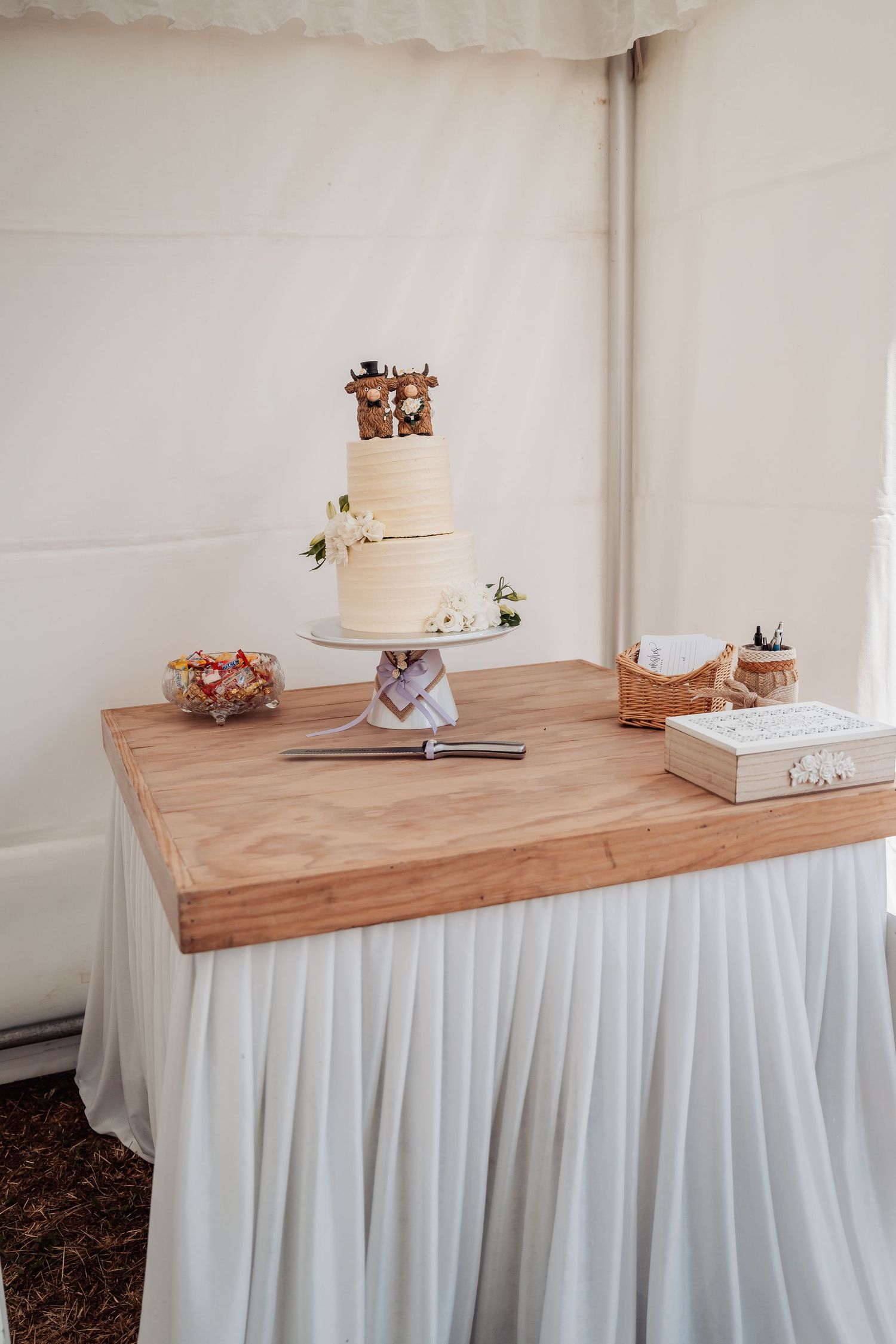 Two-tier white wedding cake with figurine topper displayed on wooden table in white marquee.