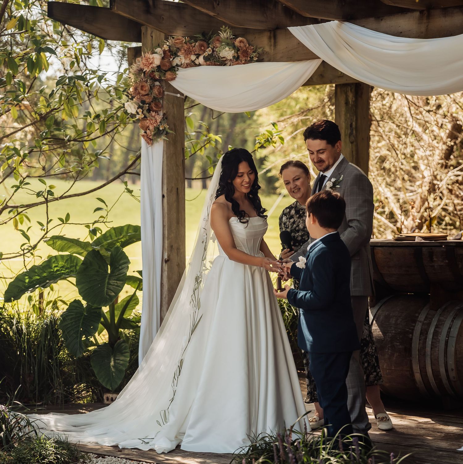 Bride and groom exchange vows under floral draped rustic arch with officiant and child witness present.