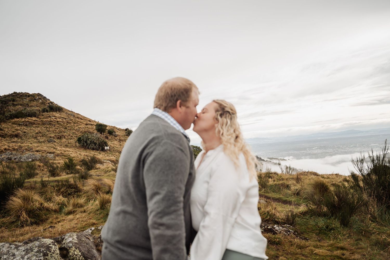 A couple in warm winter wear sharing a romantic moment on a scenic hilltop with moody cloudy skies.