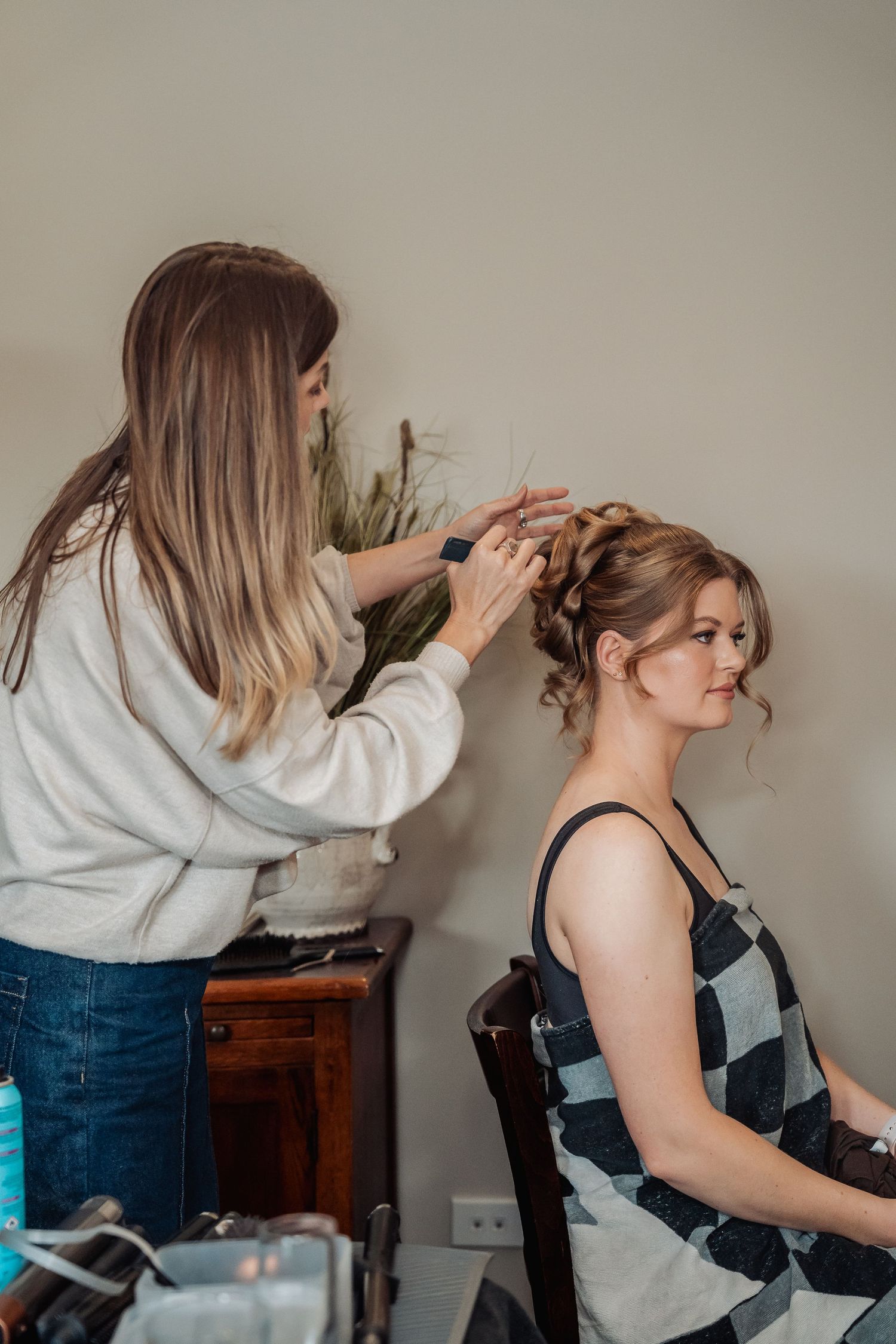 A hairstylist styles an updo while client sits in salon chair against neutral wall with wooden furniture.