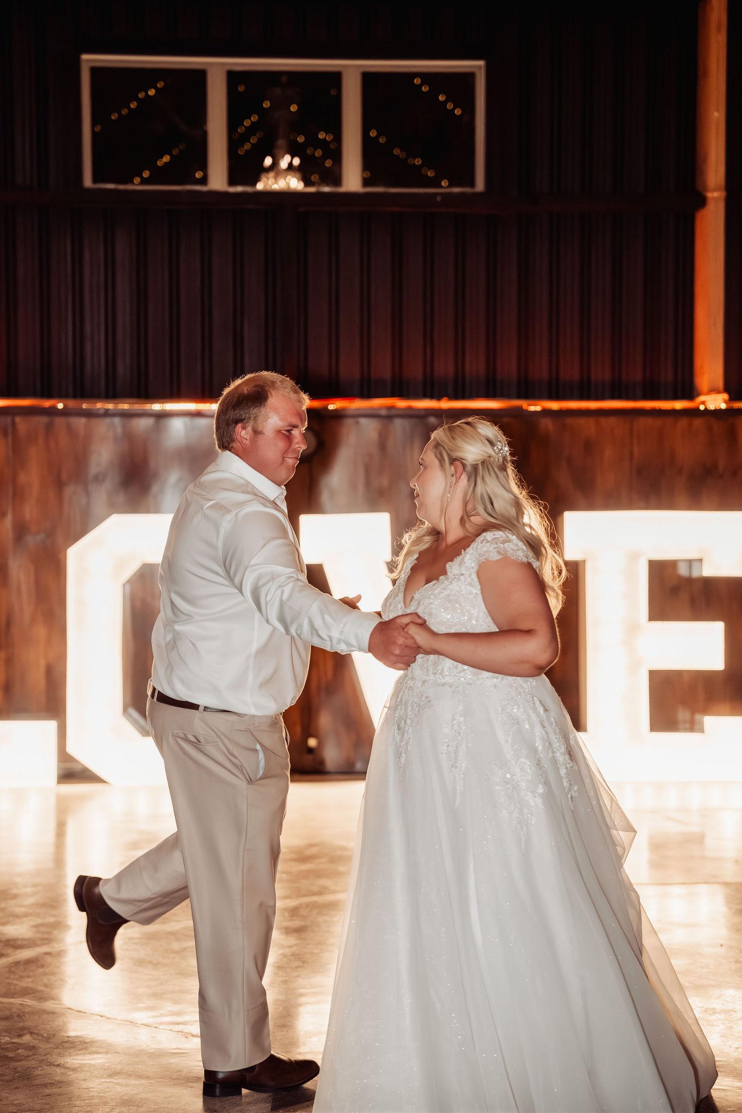 Bride and groom share a playful first dance in front of glowing LOVE letters in elegant rustic barn venue.