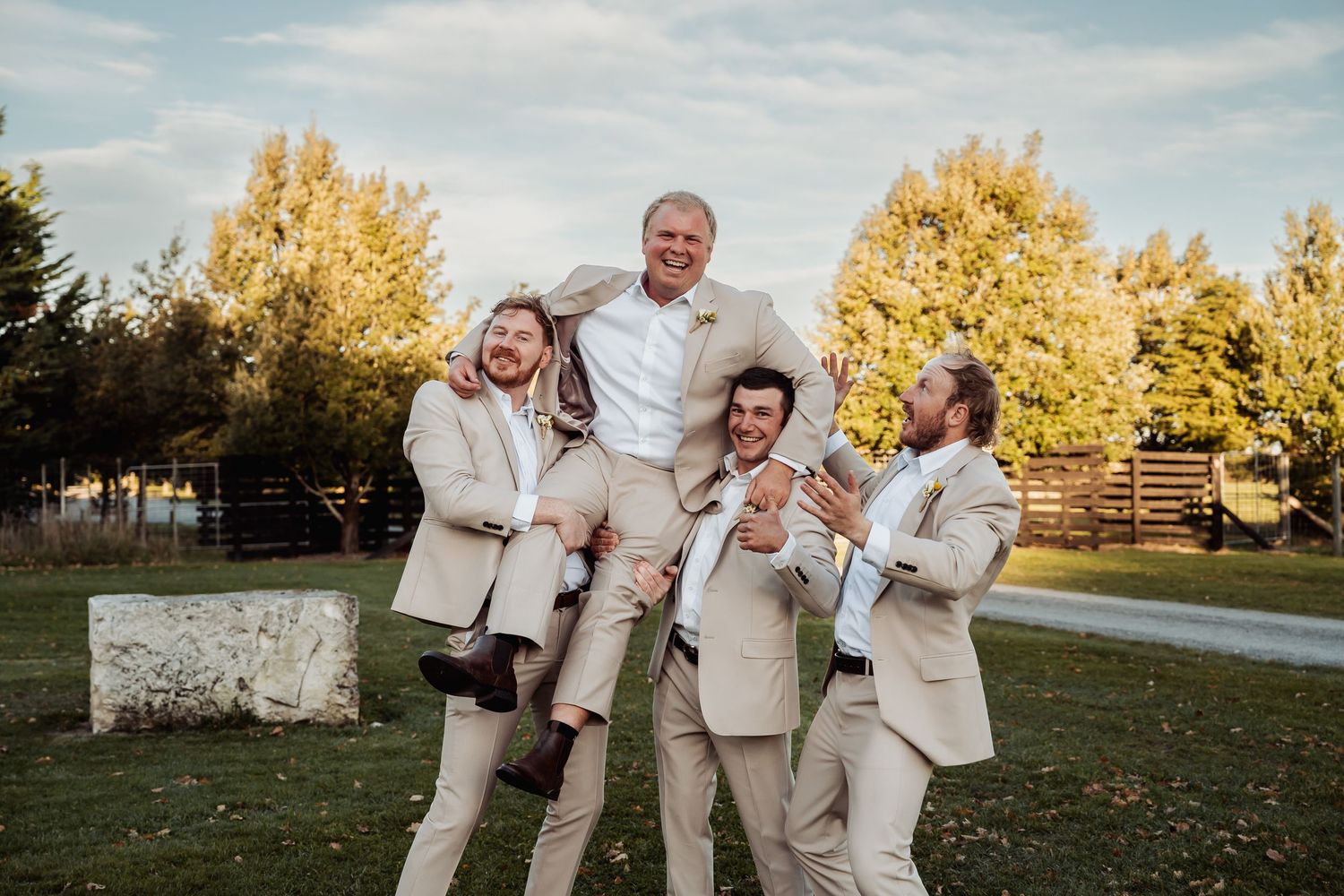 Groom in white shirt lifted by three groomsmen in matching tan suits celebrating outdoors at a wedding.