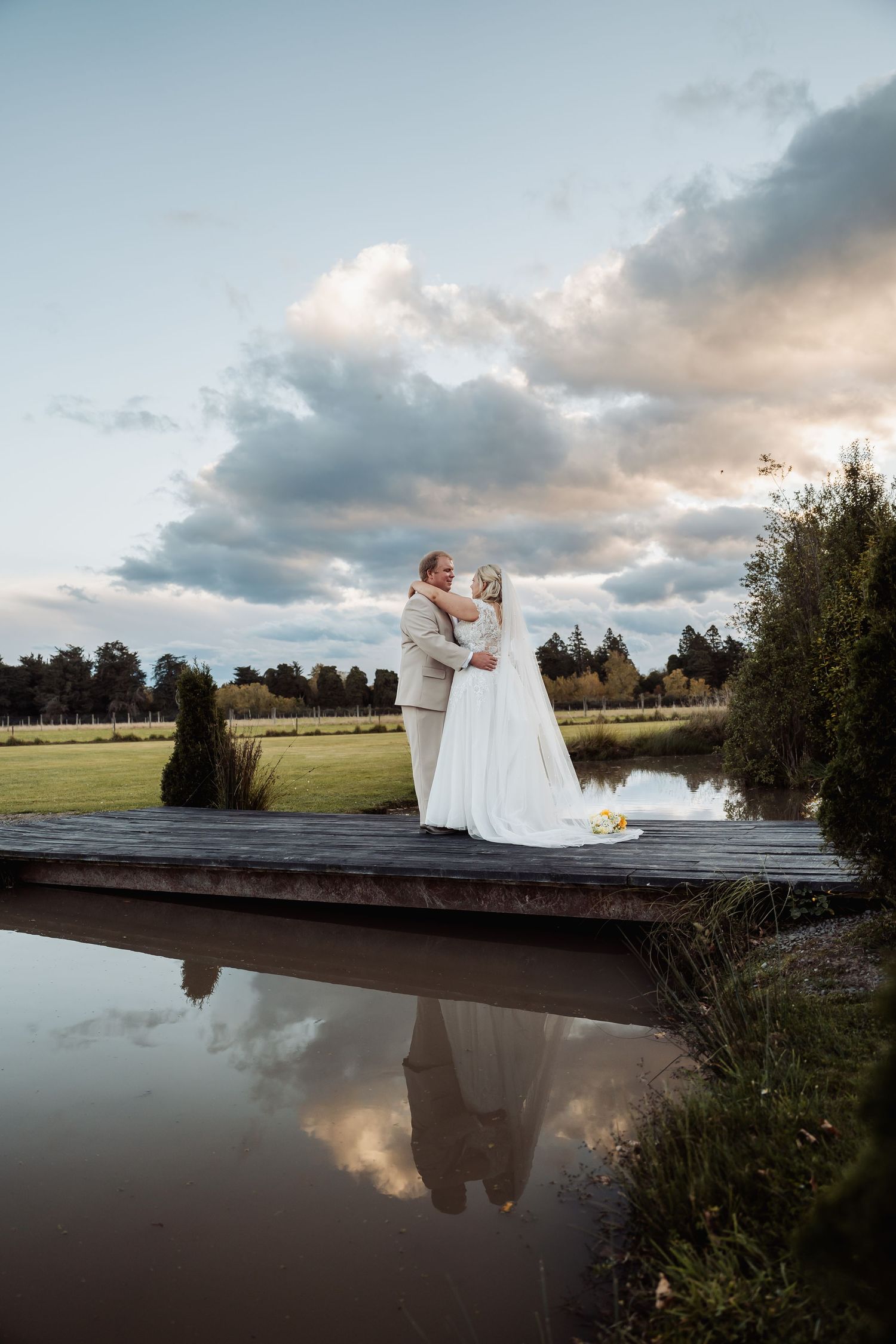 Bride and groom embrace on a wooden dock reflected in calm water under a dramatic cloudy sky at sunset.