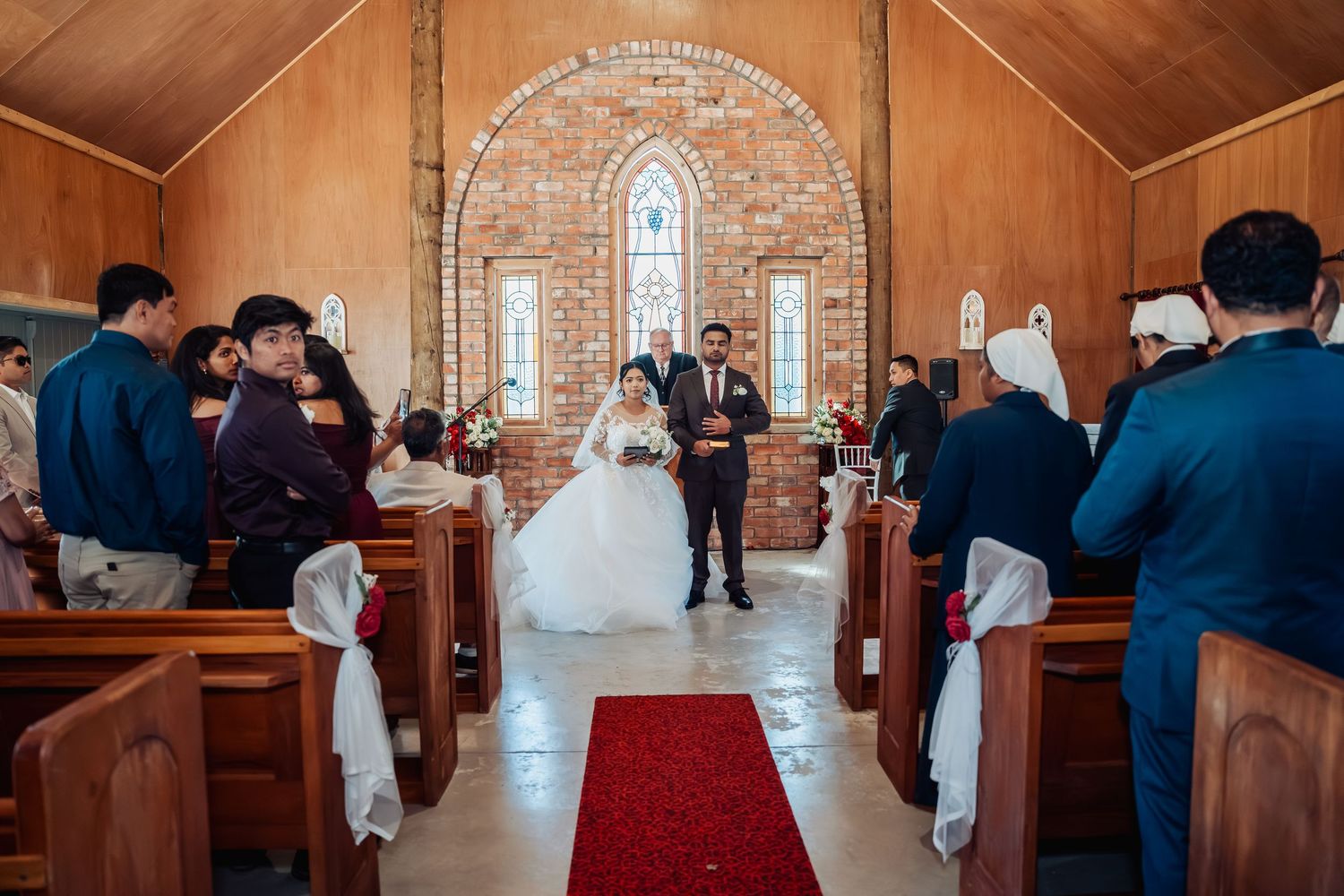 Bride and groom walk down red carpet aisle of rustic brick church surrounded by wedding guests.