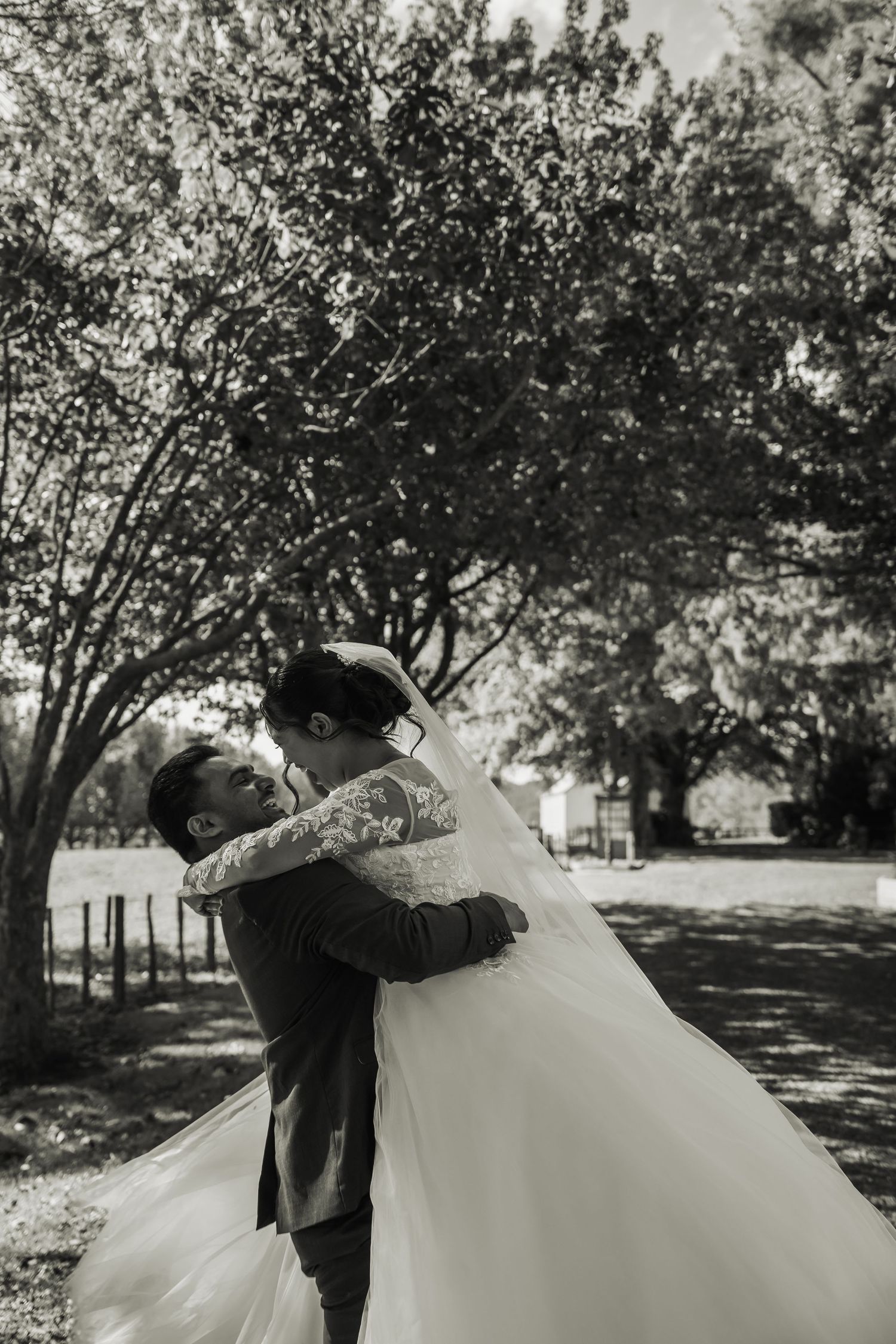 Groom lifts bride in a romantic black and white wedding photo outdoors, surrounded by lush trees.