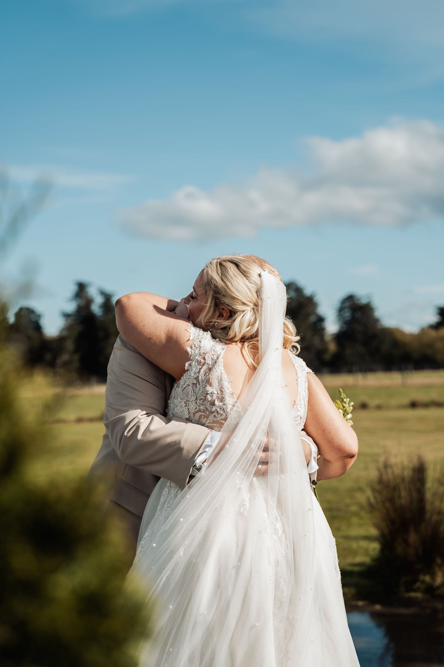 Bride and groom embrace outdoors with green fields and blue sky behind them.