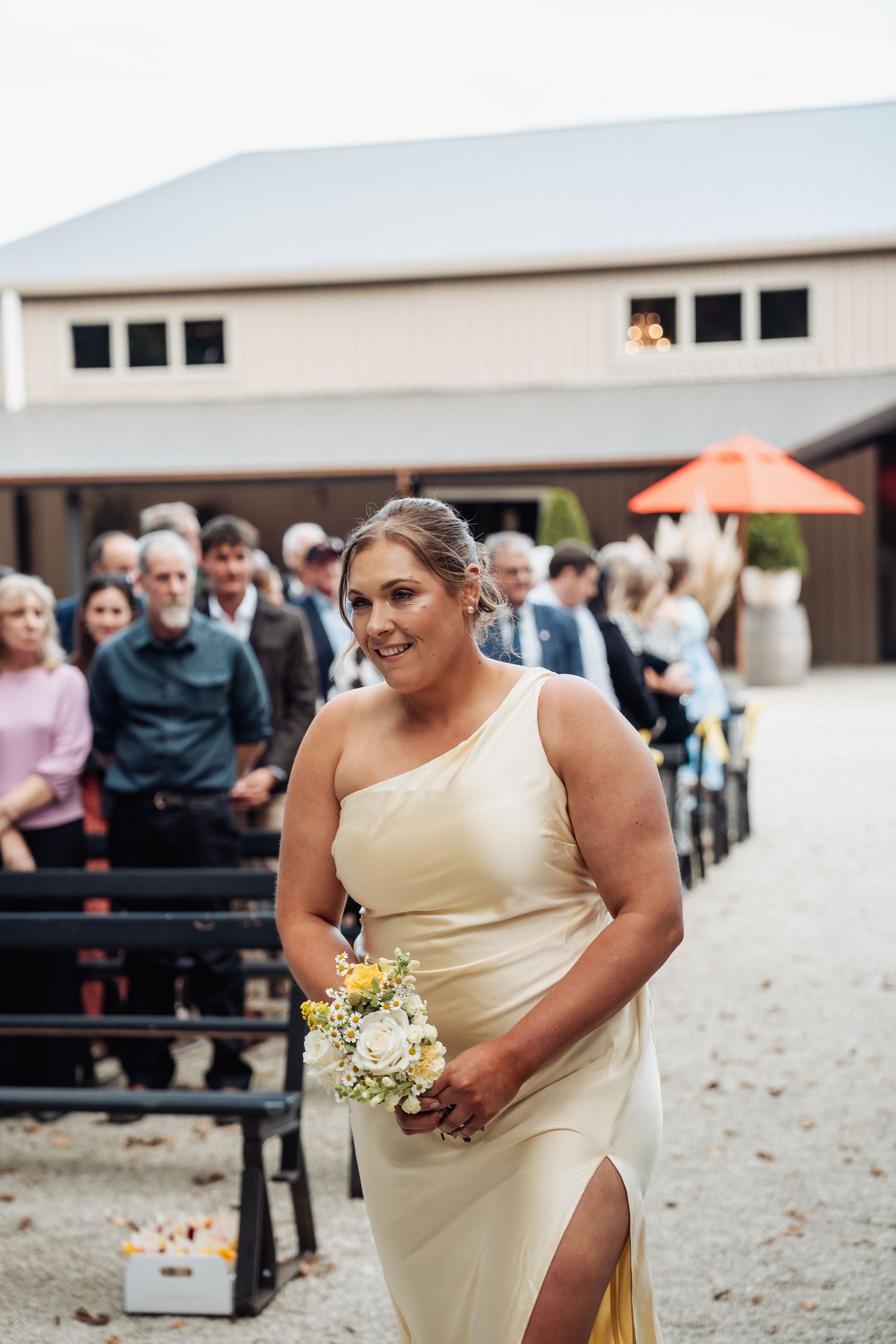 Bridesmaid in yellow one-shoulder dress walks down outdoor wedding aisle holding bouquet.