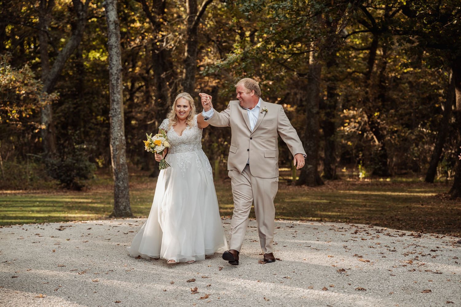 A joyful newlywed couple walks hand in hand on a gravel path surrounded by autumn trees after their ceremony.
