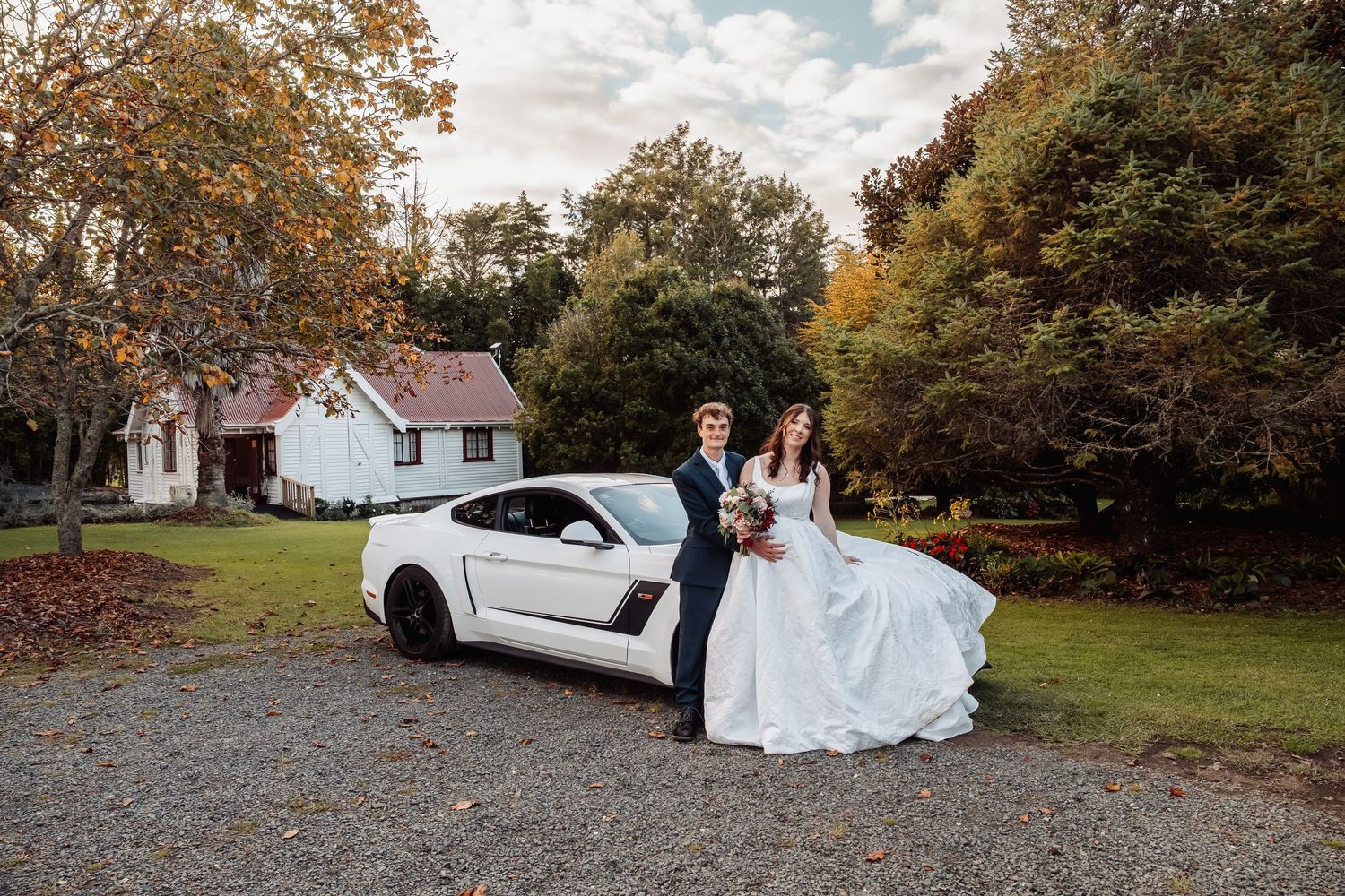 Bride and groom posing beside white Ford Mustang surrounded by colorful autumn foliage.