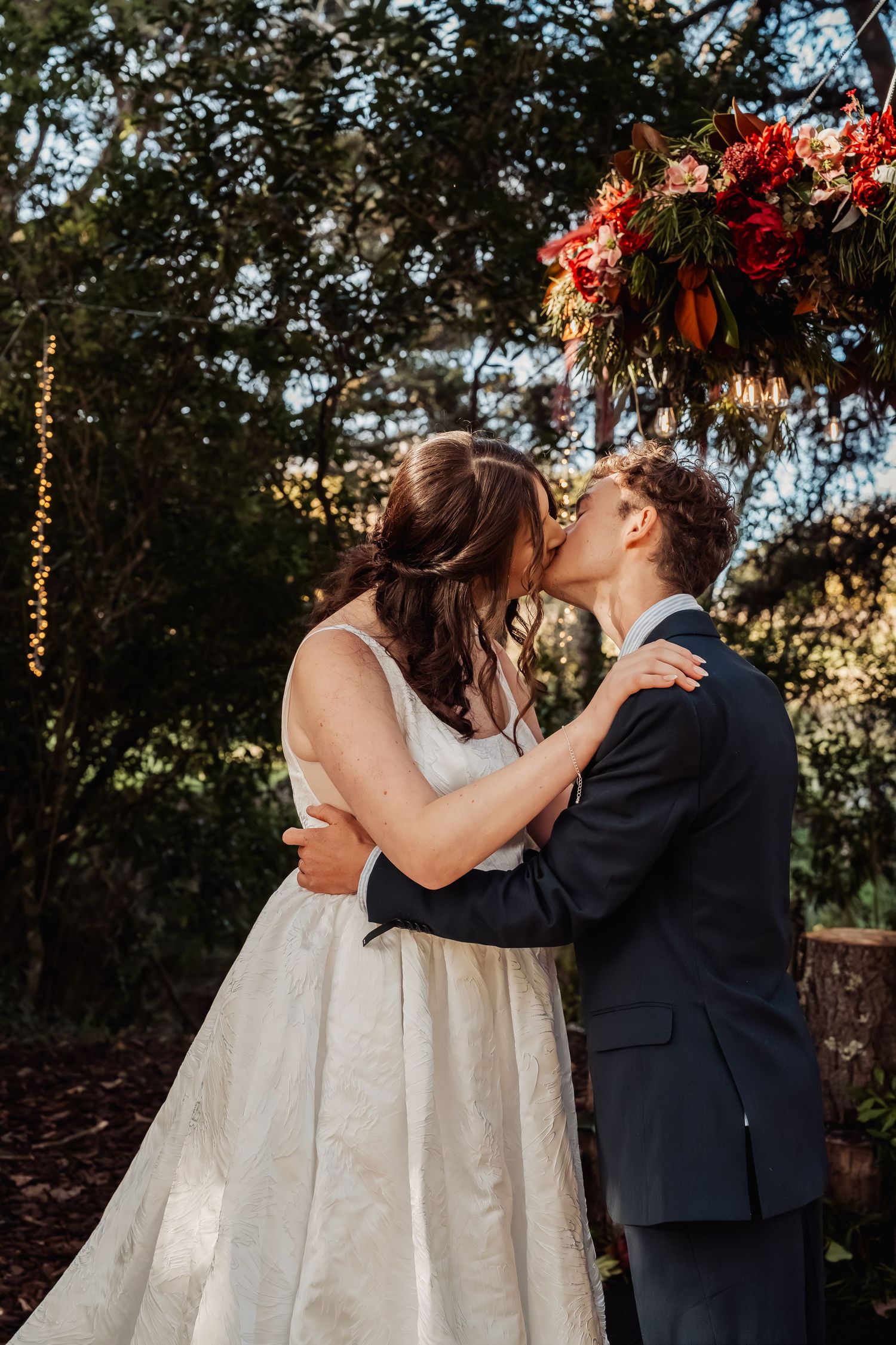 Bride and groom share first kiss under floral arch at outdoor wedding ceremony.