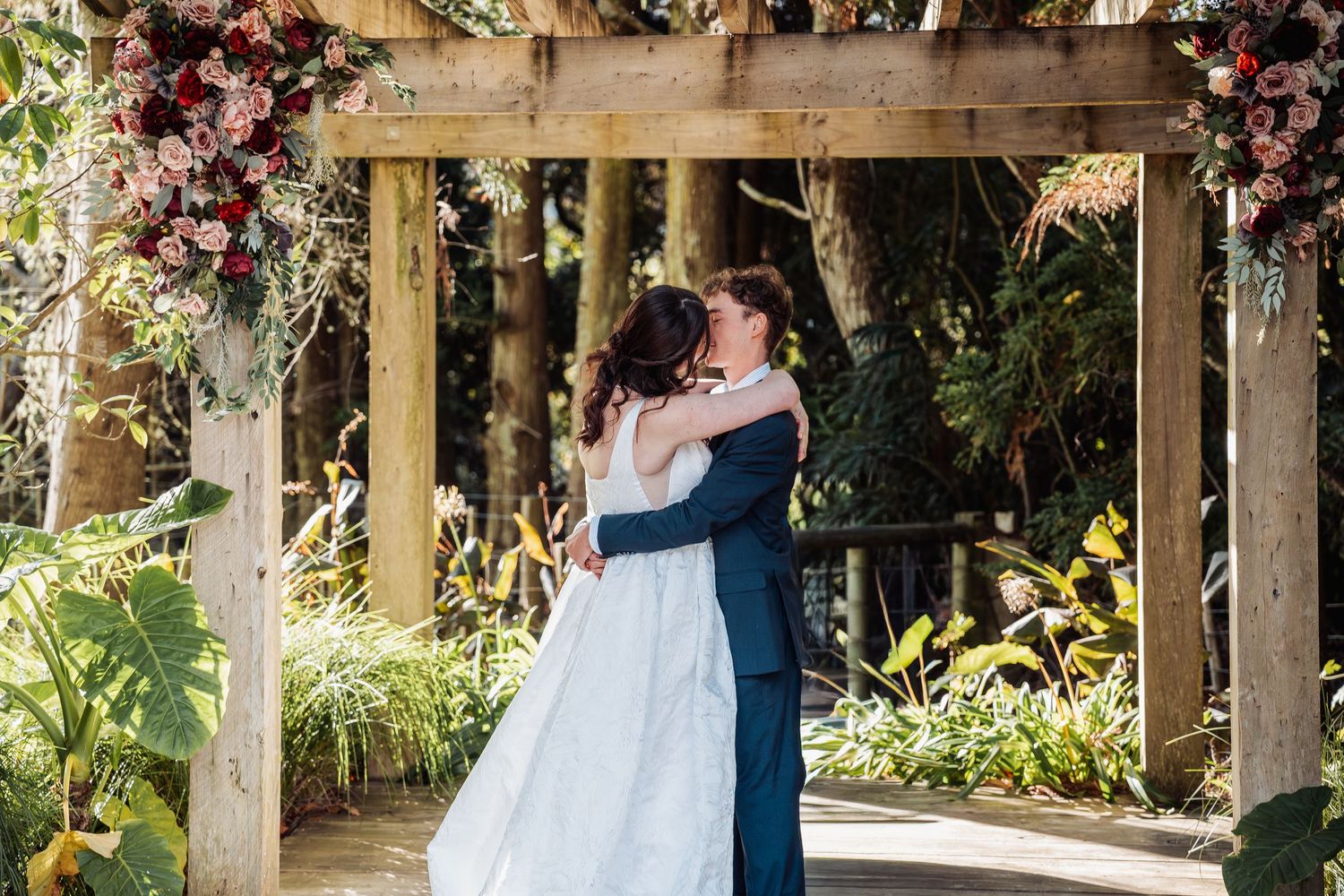 Newlyweds share first kiss under floral wooden arch surrounded by tropical garden greenery.