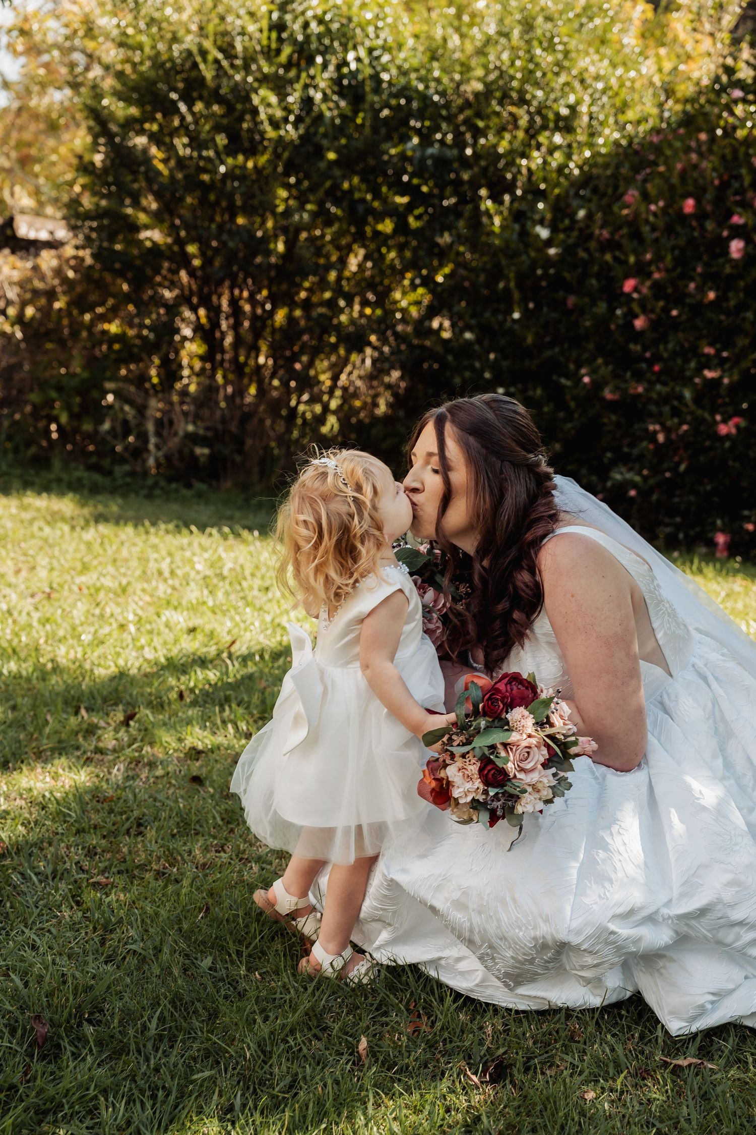 Bride in white gown crouches to kiss young flower girl in white dress on sunny garden lawn.