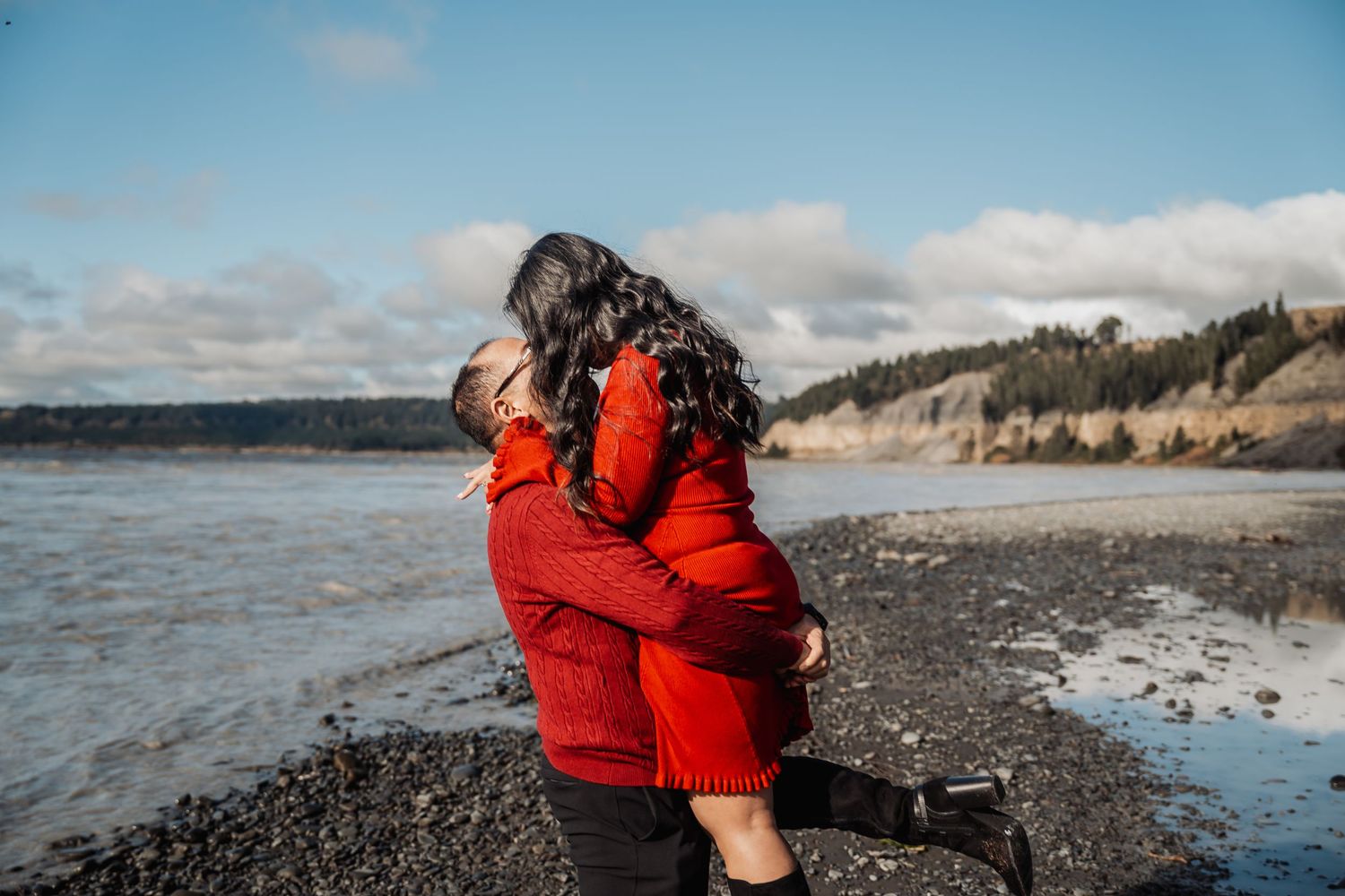 Man in red sweater lifts woman in red dress in a joyful embrace on a rocky beach.